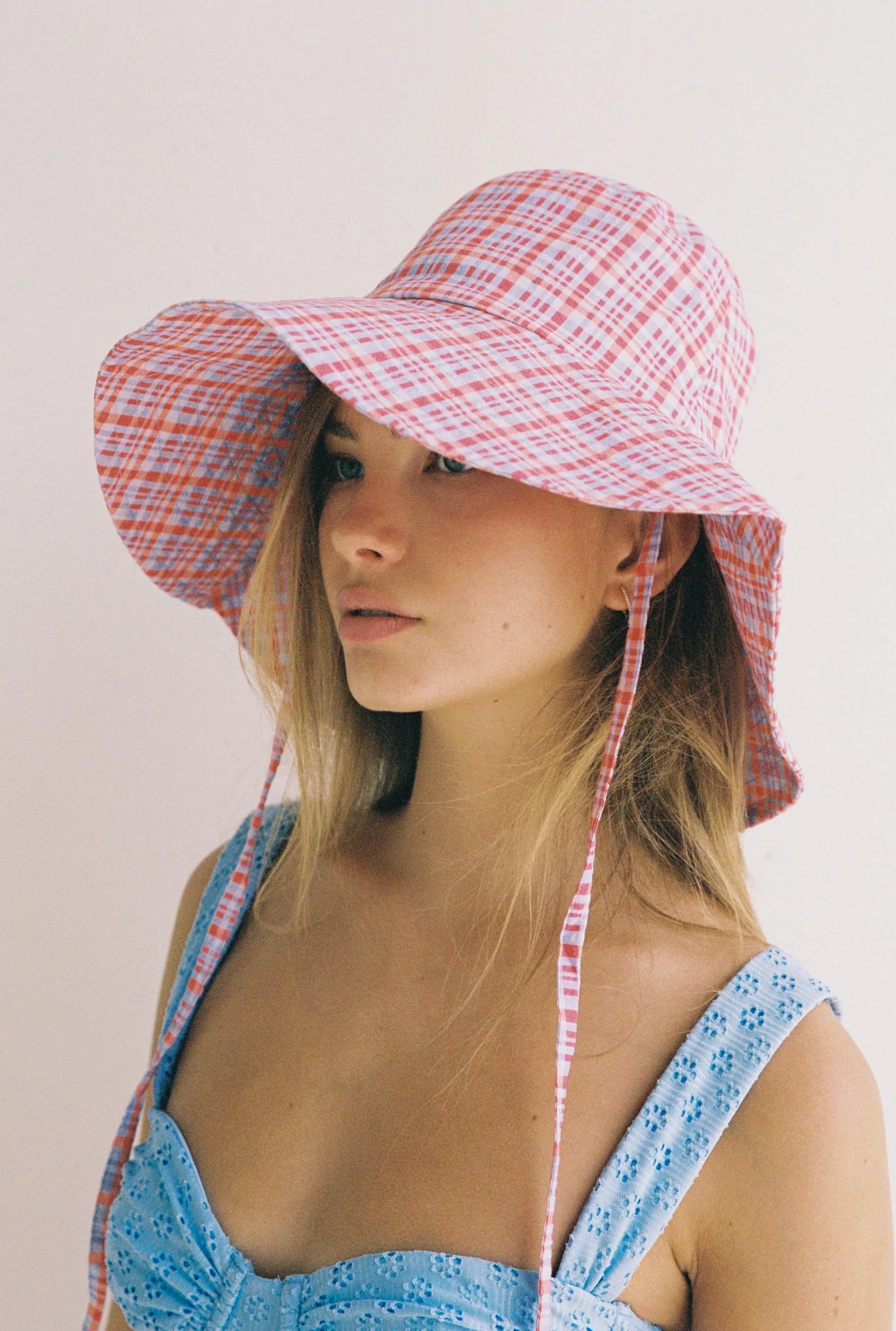 A person dons the JUNI ~ Picnic Check Sunhat, a unique design with a wide brim in red and pink stripes, paired with a blue eyelet dress. The hat's chin straps drape casually against the plain white background.