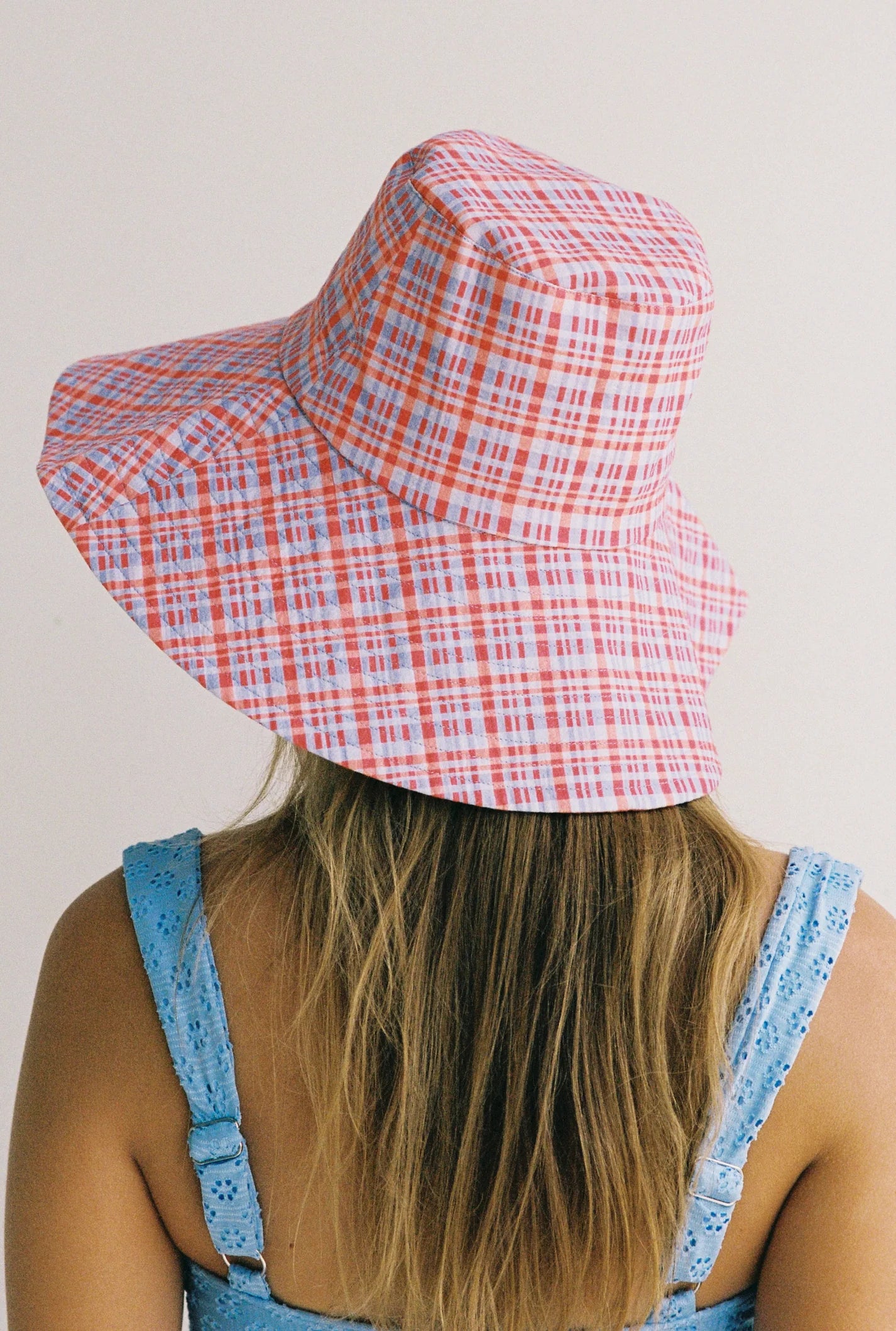 A person with long hair faces away from the camera, wearing the JUNI ~ Picnic Check Sunhat made from pink and blue seersucker fabric, paired with a light blue eyelet top against a plain background.