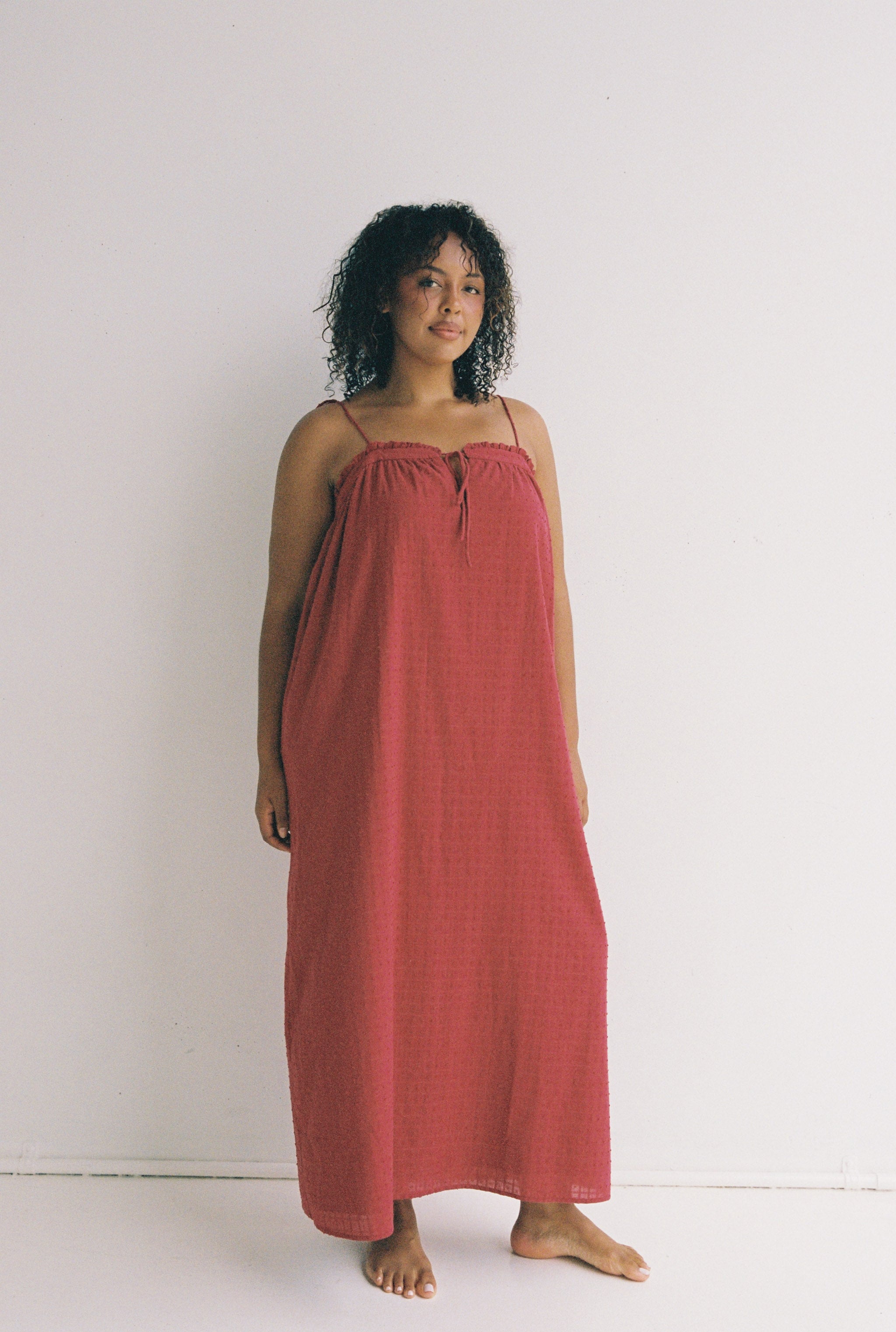 A person with curly hair stands barefoot against a plain white background, wearing the Marlow Maxi Dress Merlot by JUNI, a long, sleeveless red dress with adjustable shoulder ties and functional pockets.