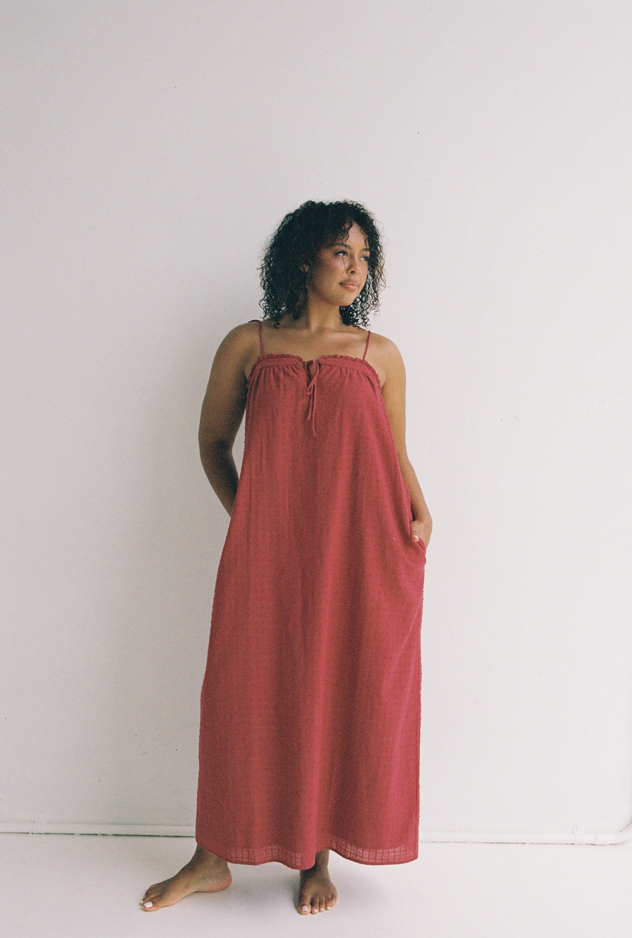 A person with curly hair is standing barefoot against a plain white background, wearing the Marlow Maxi Dress Merlot by JUNI. The dress is long, sleeveless, features thin straps and functional pockets. With one hand in a pocket, they are looking slightly to the side.