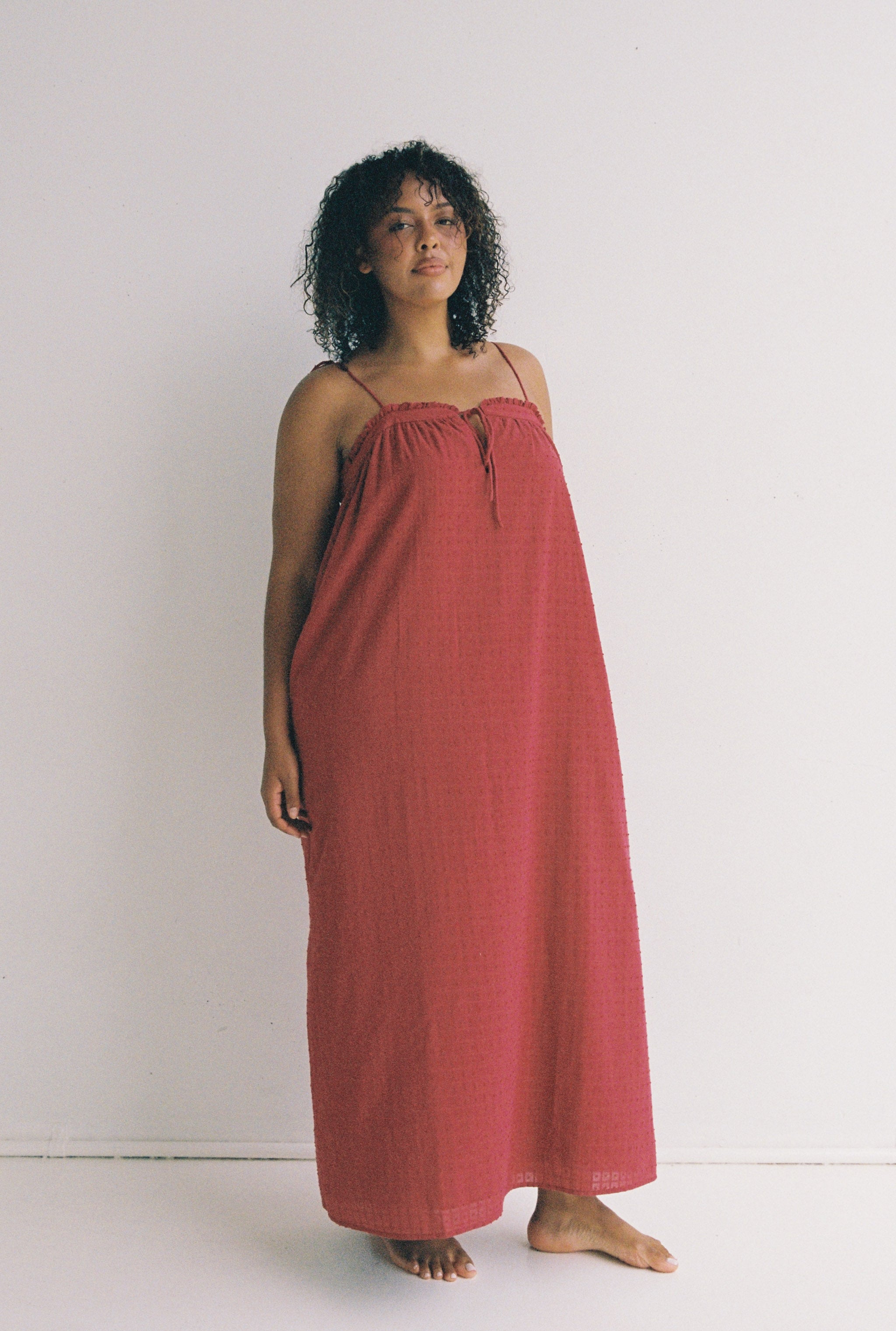 A person with curly hair stands barefoot in the Marlow Maxi Dress Merlot by JUNI, a red, sleeveless dress that includes functional pockets. They pose against a plain white background, looking at the camera with a neutral expression.