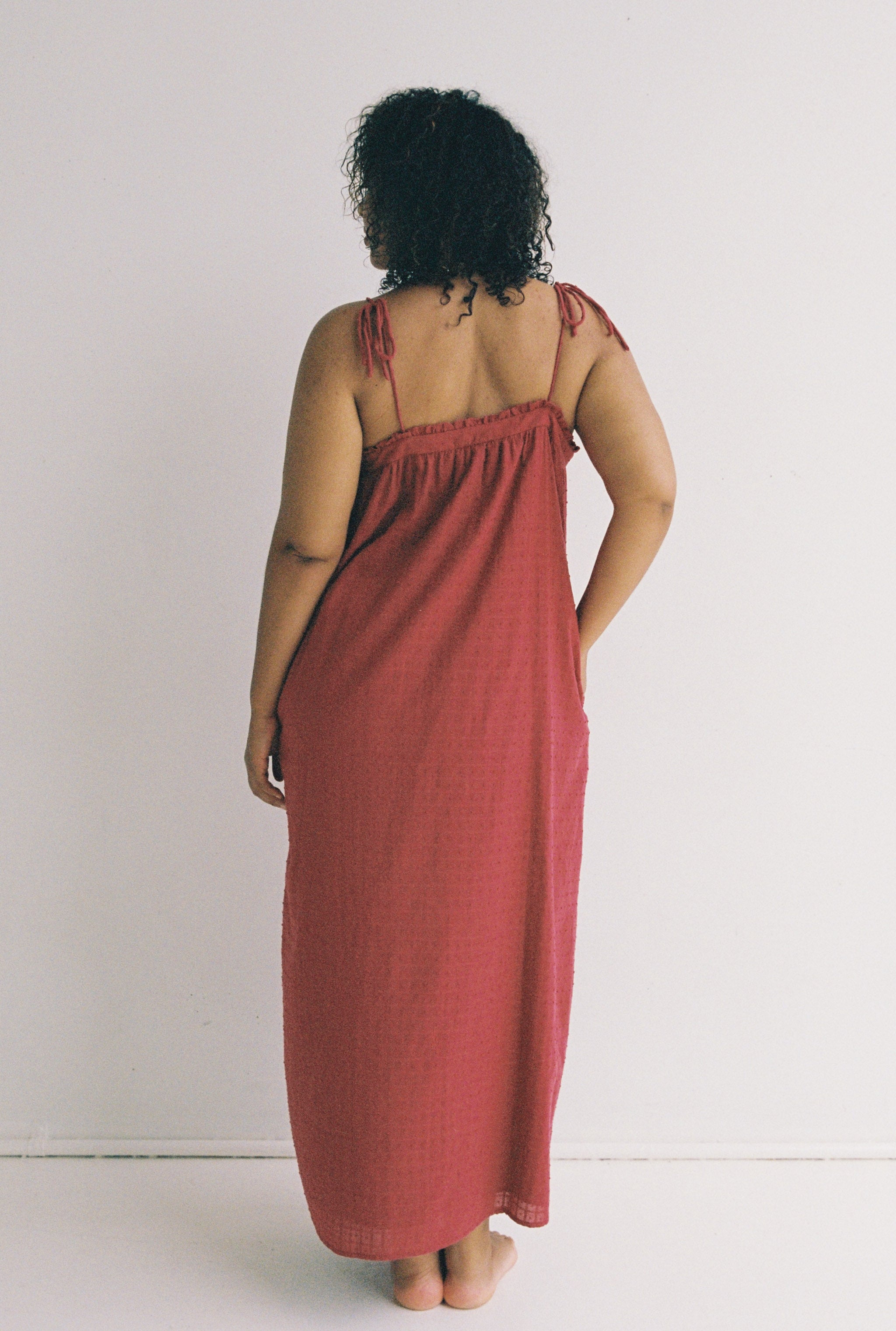 A person with curly hair stands barefoot, facing away from the camera, wearing the Marlow Maxi Dress Merlot by JUNI, a long, sleeveless red dress featuring adjustable shoulder ties. Against a plain white wall backdrop, this elegant piece exemplifies style with consciousness and is ethically made.