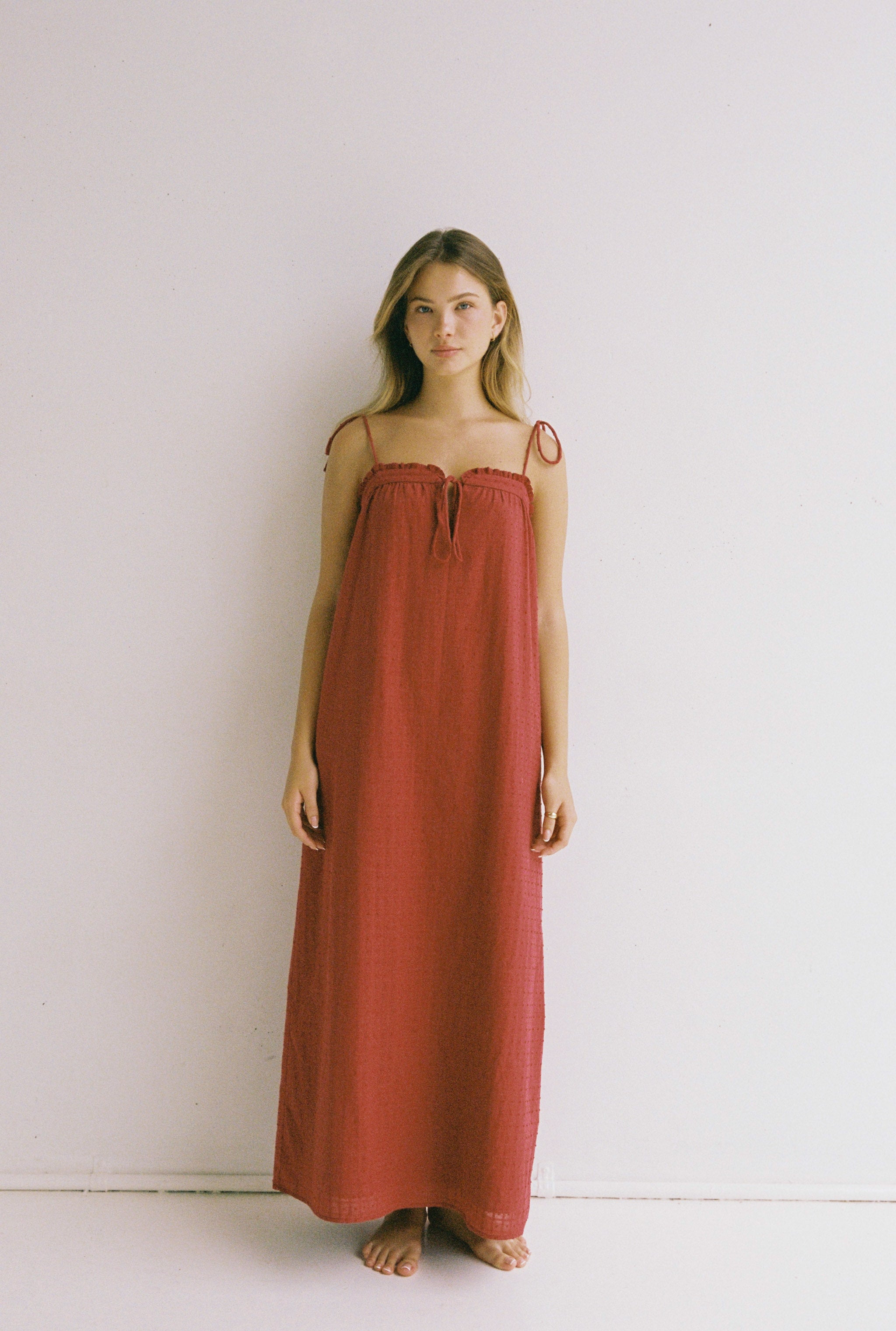 A person with long hair stands barefoot against a plain white background, wearing the Marlow Maxi Dress in Merlot by JUNI, featuring an ethically made design with adjustable shoulder ties and thin straps.