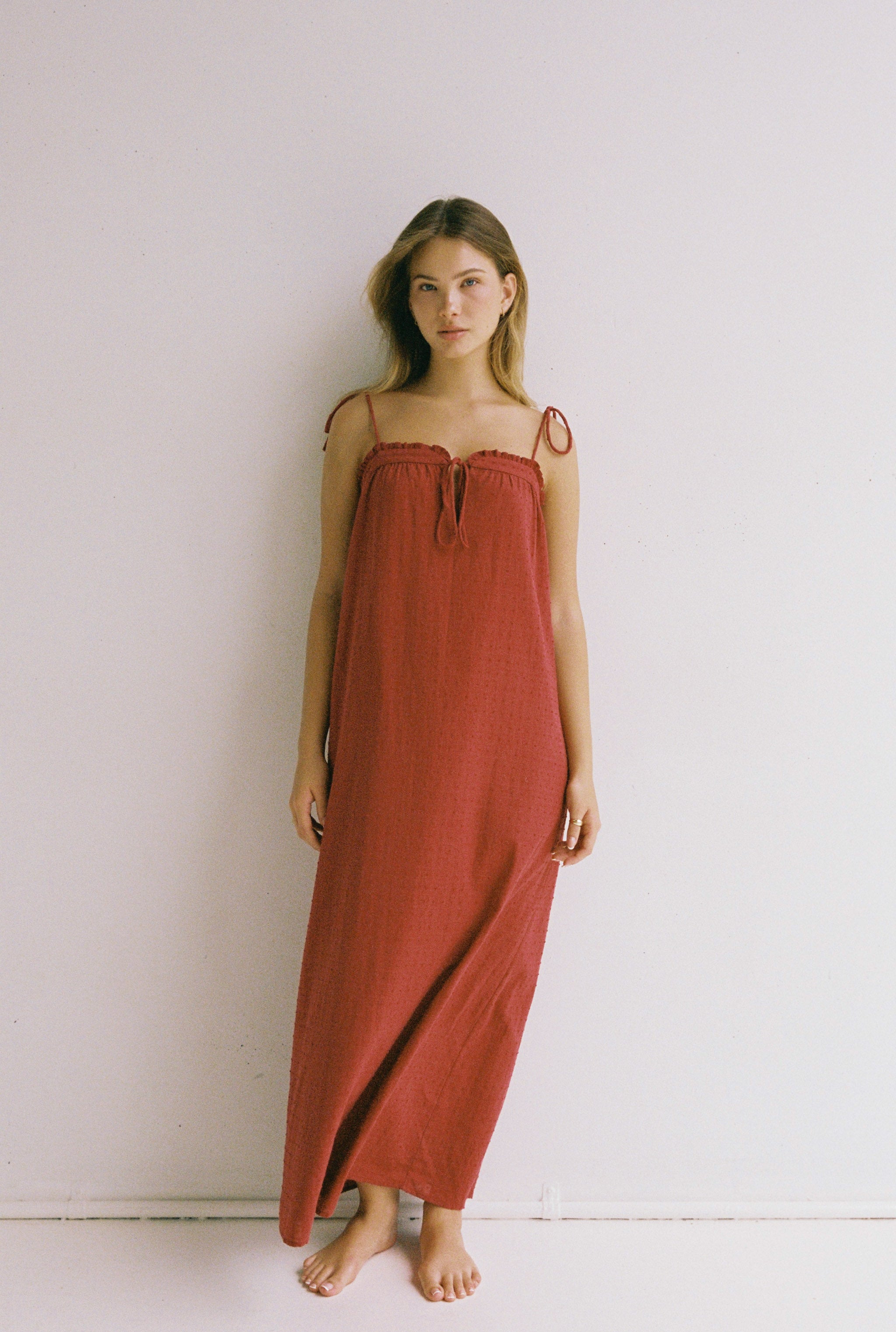 A woman stands against a white wall, wearing the Marlow Maxi Dress in Merlot by JUNI. The dress is long and sleeveless, featuring adjustable shoulder ties. She is barefoot and gazes slightly to the side with a relaxed expression, embodying the essence of an ethically crafted piece that seamlessly blends style and conscience.