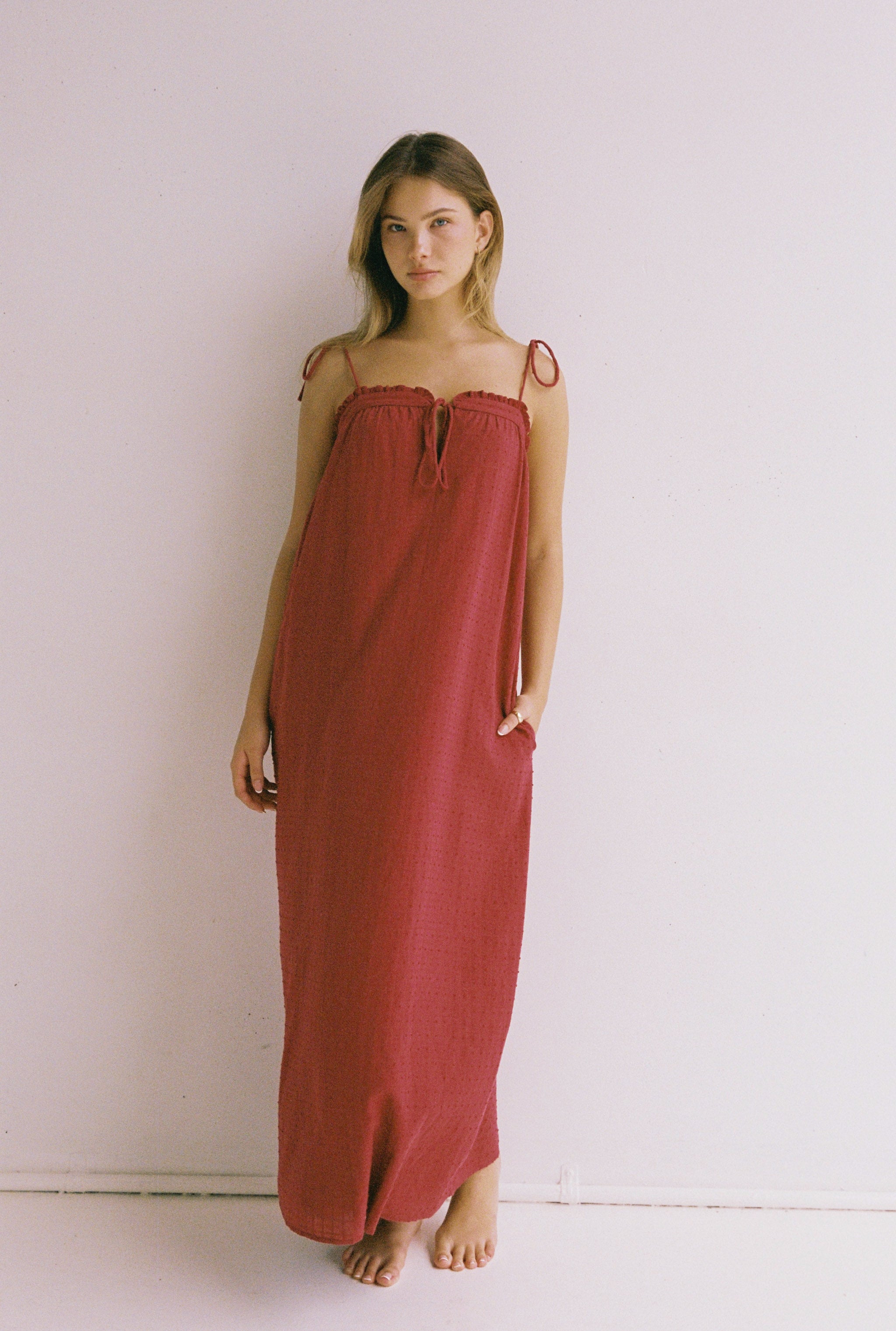 A woman stands barefoot against a plain white wall, wearing the Marlow Maxi Dress in Merlot by JUNI. The long, sleeveless dress features adjustable shoulder ties, and she rests her hands casually in the functional pockets while gazing directly at the camera.