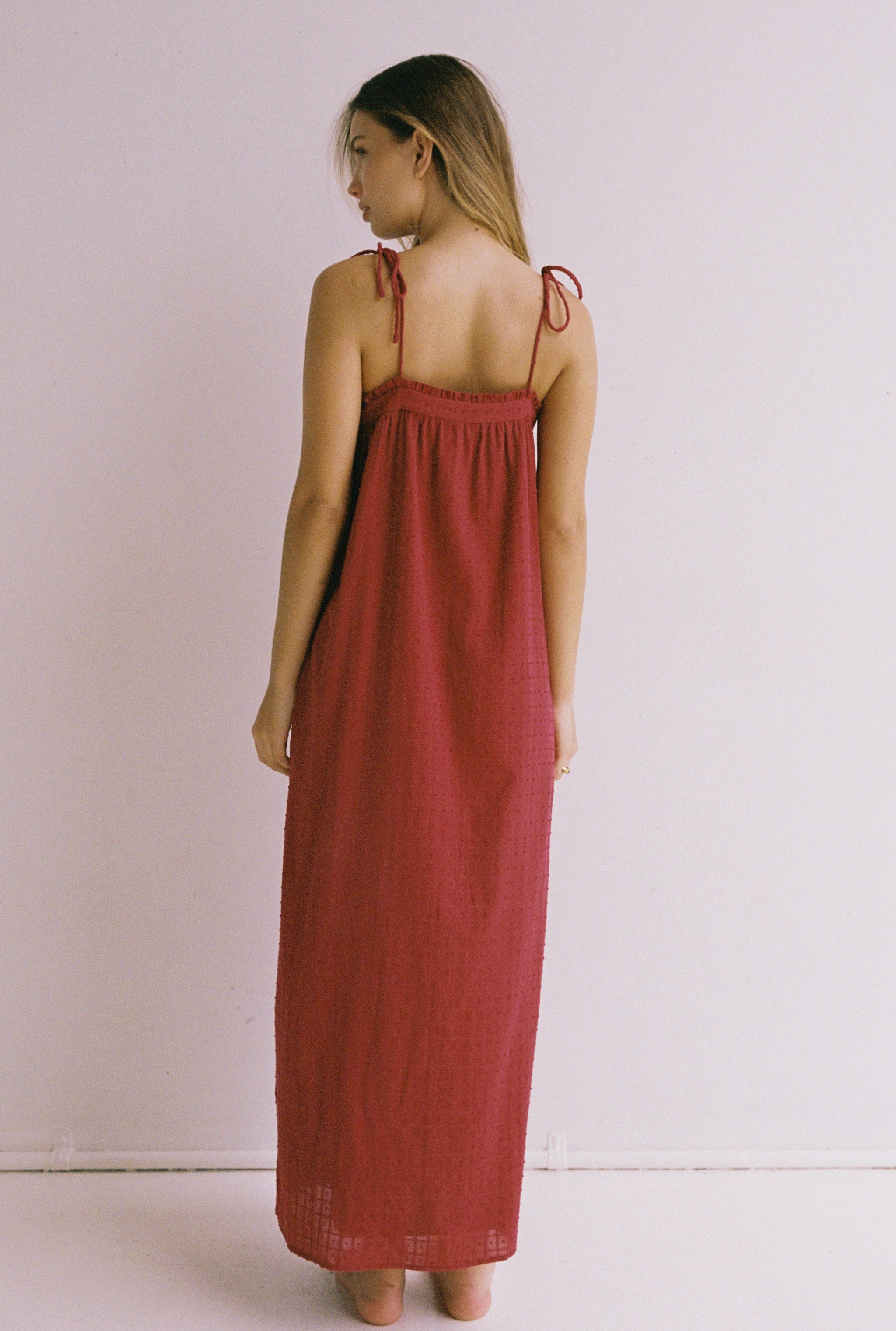 A woman stands with her back to the camera, showcasing the Marlow Maxi Dress Merlot by JUNI. The dress features a long, flowing design with adjustable shoulder ties and is set against a plain, light-colored background. This ethically made piece blends elegance and comfort seamlessly.