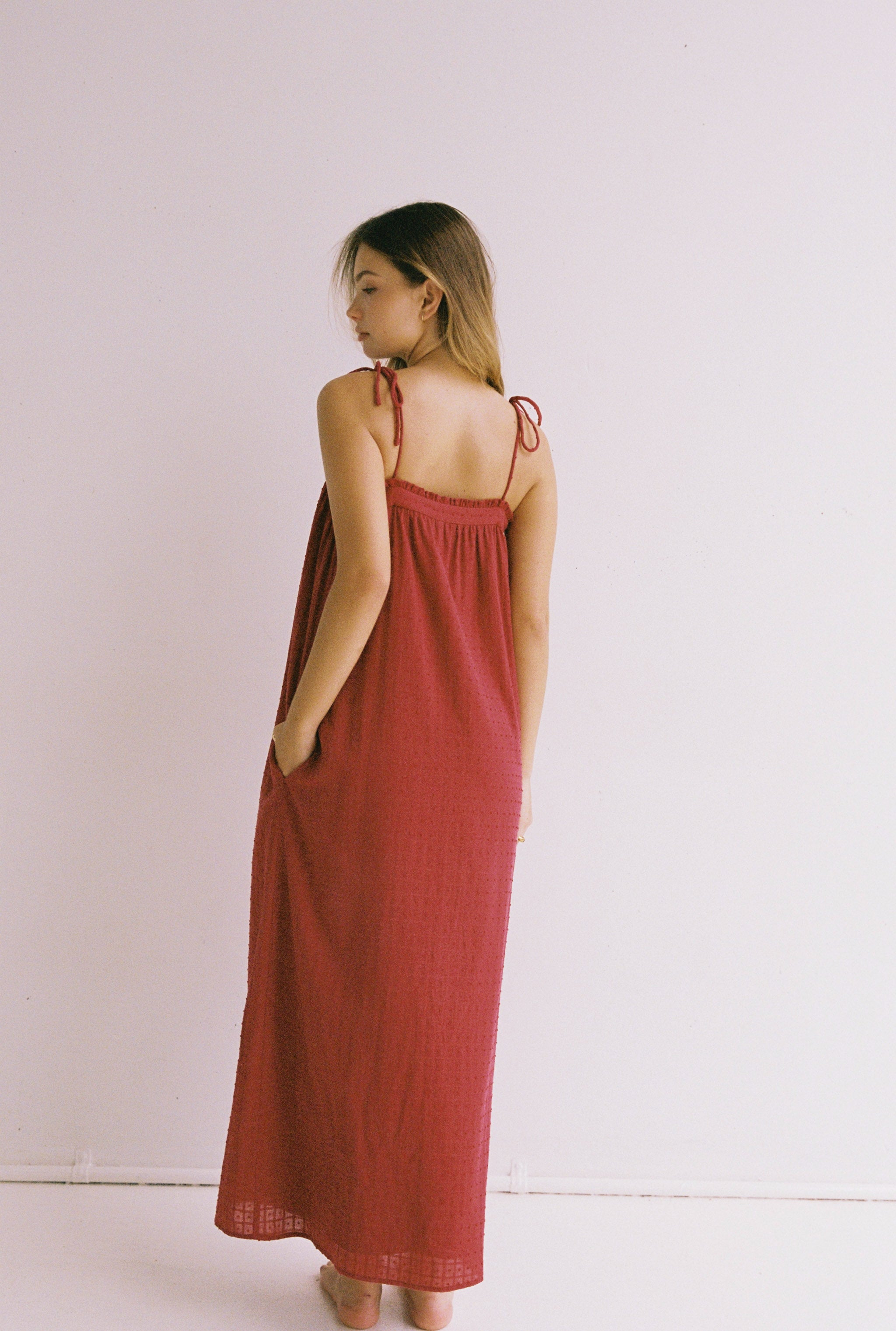 A woman wearing the Marlow Maxi Dress Merlot by JUNI stands barefoot, with adjustable shoulder ties and her light brown hair flowing freely. She poses against a plain white background, perfectly embodying the essence of ethically made fashion.