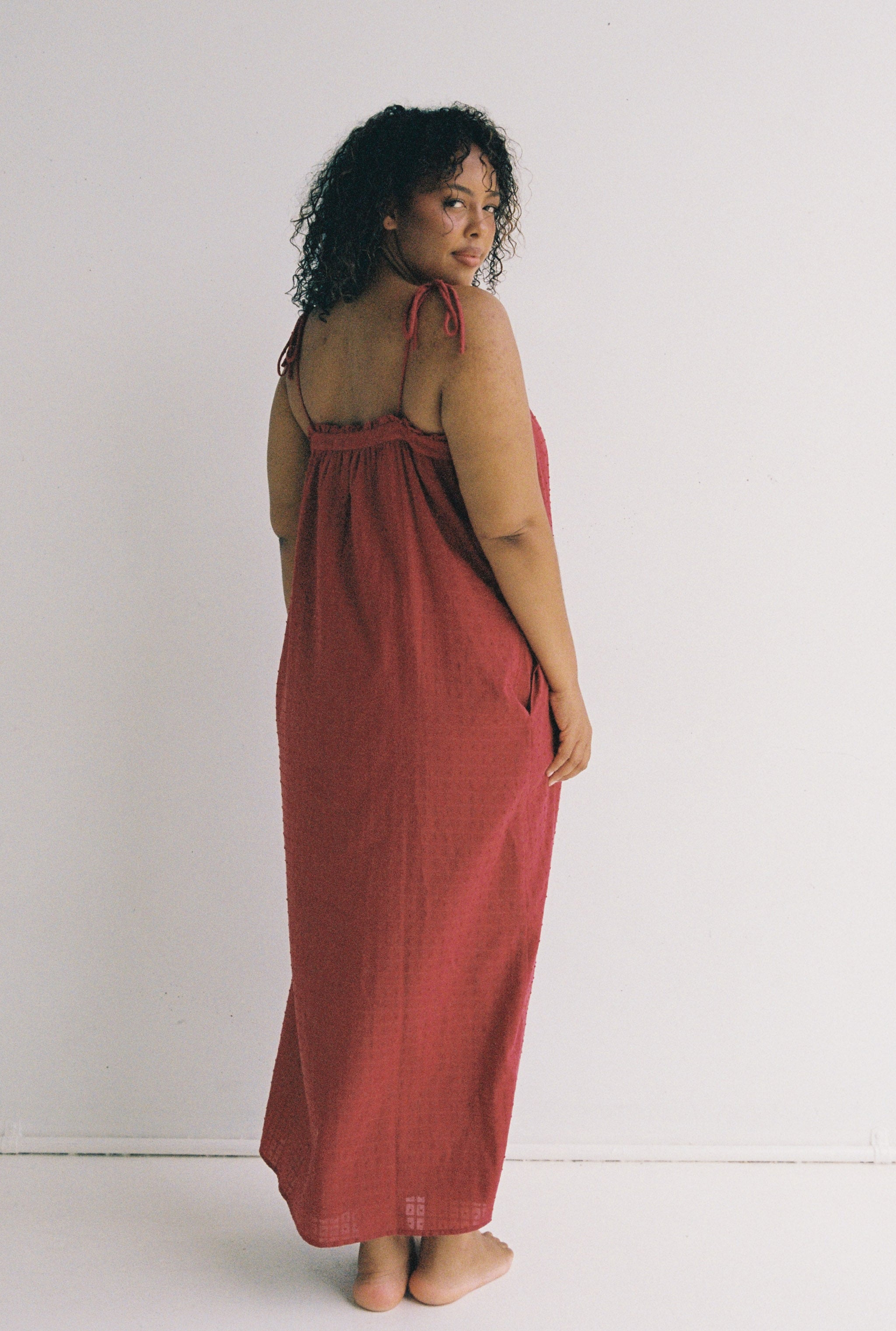 A person with curly hair stands barefoot against a plain white background, wearing the Marlow Maxi Dress Merlot by JUNI. They are facing slightly to the side, looking at the camera over their shoulder.