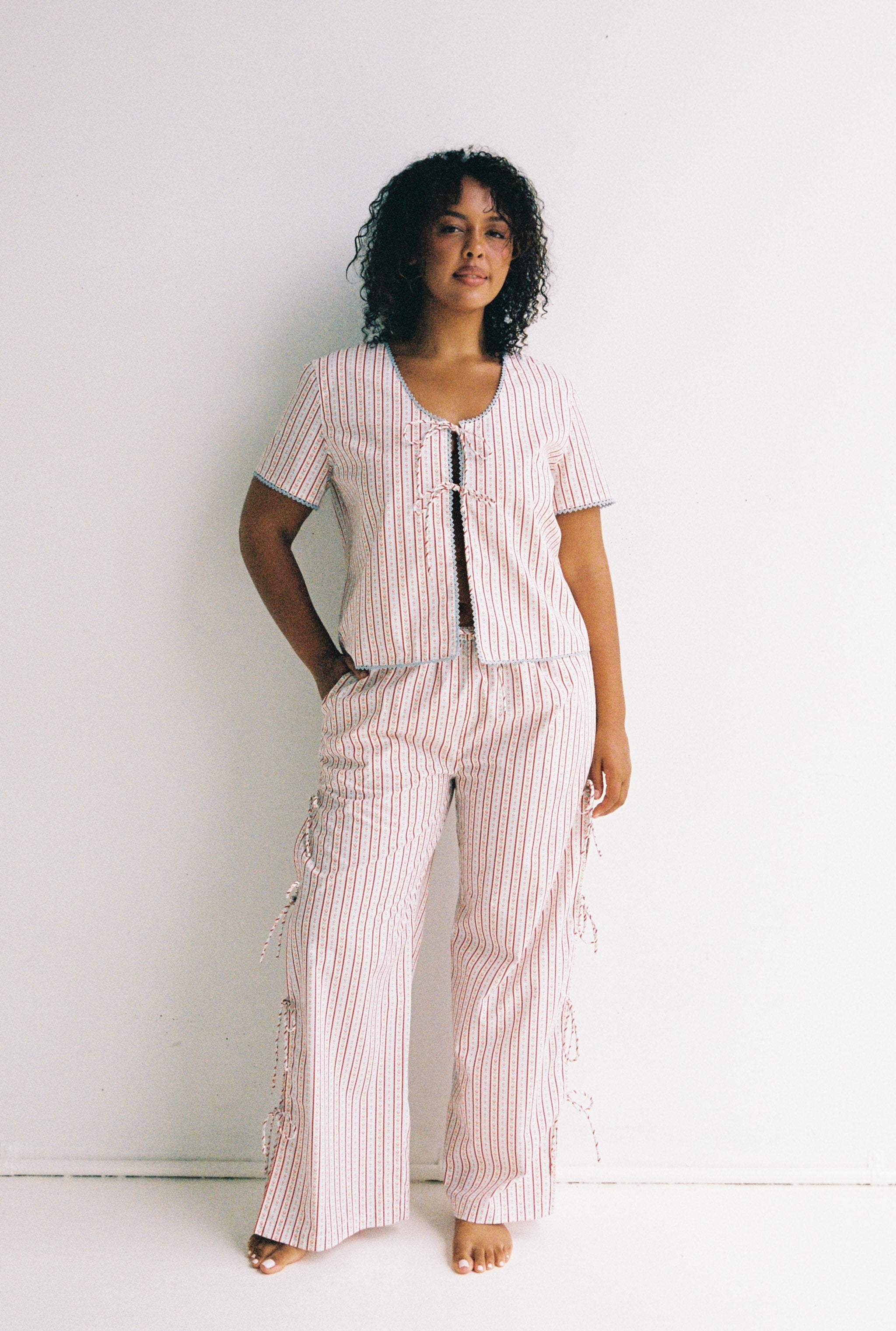 A person with curly hair stands against a white background, wearing a coordinated ensemble featuring the Poppy Blouse by JUNI, crafted from cotton poplin. The short-sleeved top and pants exude comfort, with one hand in their pocket, conveying a relaxed demeanor.