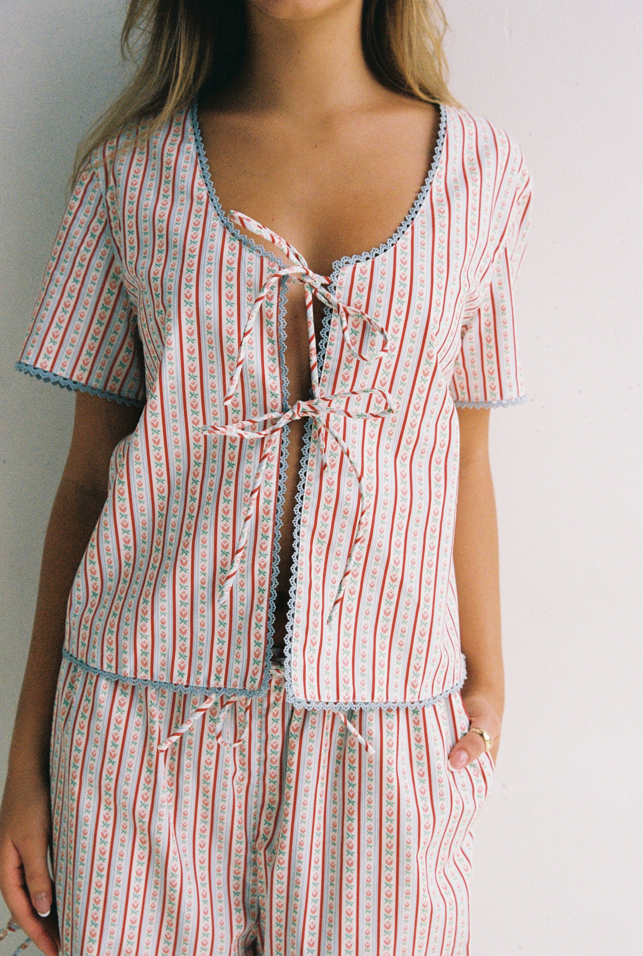 A person showcases the Poppy Blouse from JUNI, a striped outfit with red and blue vertical patterns, featuring a short-sleeve design with lace trim, a scooped neckline, and adorned with bows. The ensemble is completed by pocketed pants as they pose against a plain, light-colored background.