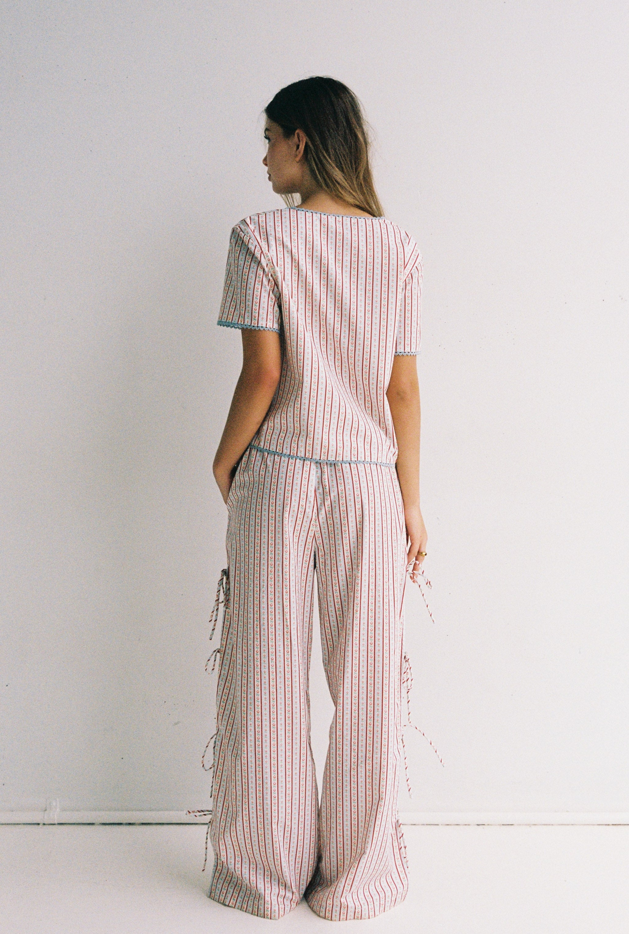 A person is wearing a matching outfit featuring the Poppy Blouse by JUNI, which has a striped pattern, short sleeves, and a scooped neckline. They pair it with wide-legged cotton poplin pants that have side ties. The person stands against a plain white background and faces slightly away from the camera.