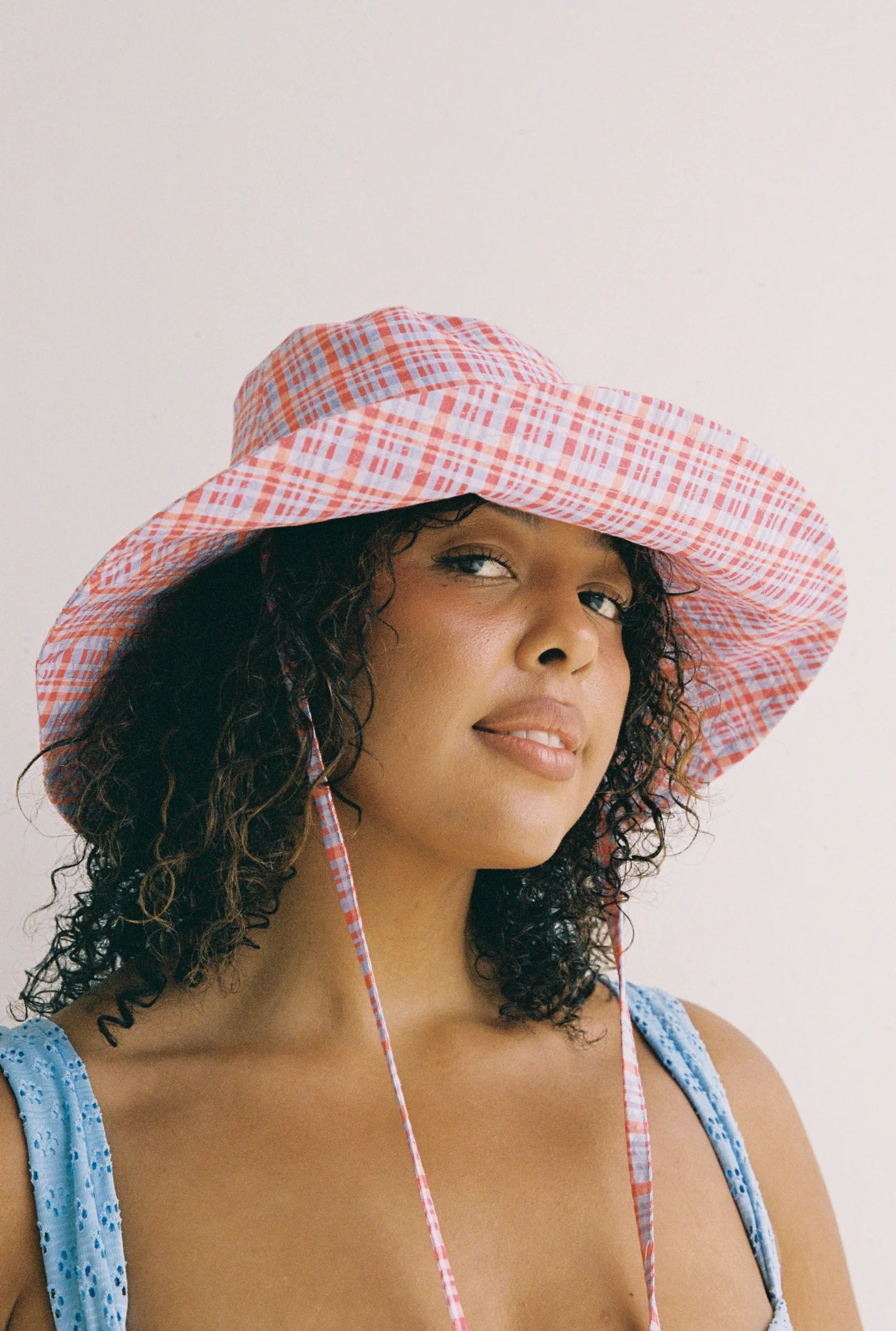 A person with curly hair sports the JUNI ~ Picnic Check Sunhat, a pink and blue plaid bucket hat from JUNI, crafted from seersucker fabric. They are also wearing a light blue sleeveless top and are looking at the camera against a plain background.