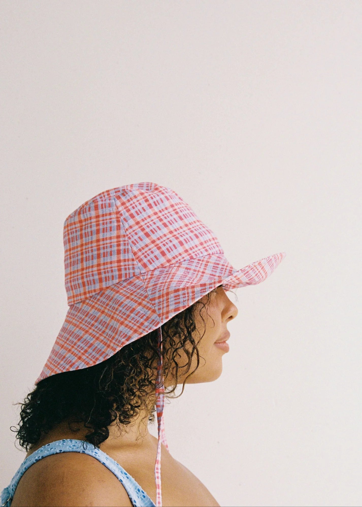 A person with curly hair wears the JUNI ~ Picnic Check Sunhat by JUNI, which features an exclusive red and white plaid print. The wide brim casts a shadow on their face as they stand against a plain, light-colored background, dressed in a blue thin-strapped top.