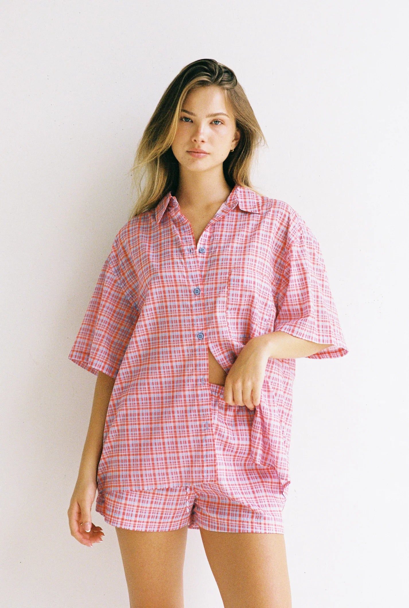 A person with long hair poses against a plain background wearing the JUNI Watson Shirt, an oversized short-sleeve button-up with matching shorts in pink plaid. Made from 100% cotton and ethically produced, they have one hand partially in a pocket.