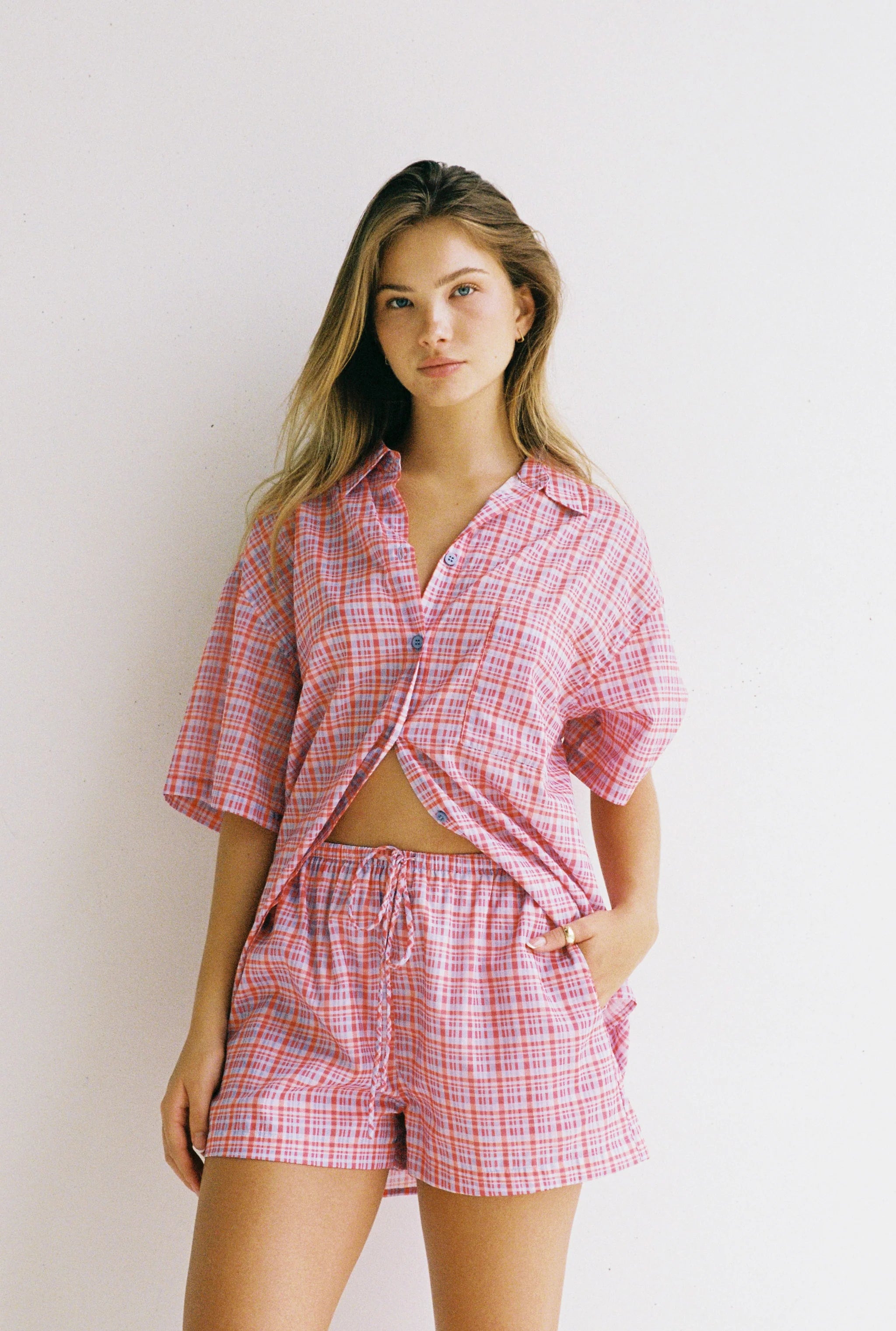 A person with long hair and a neutral expression stands against a plain background wearing JUNI's Dylan Short, paired with a relaxed fit pink and white plaid button-up shirt, partially tucked in.