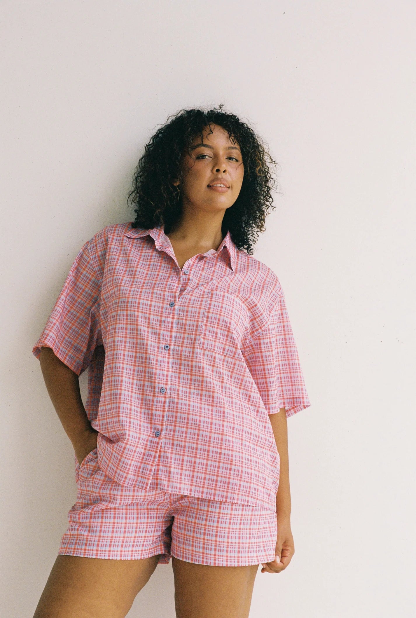 With curly hair, a person wears the JUNI Watson Shirt—an ethically made, oversized pink and white plaid short-sleeve button-up with matching shorts. Made from 100% cotton, they stand against a plain white background with one hand in a pocket.