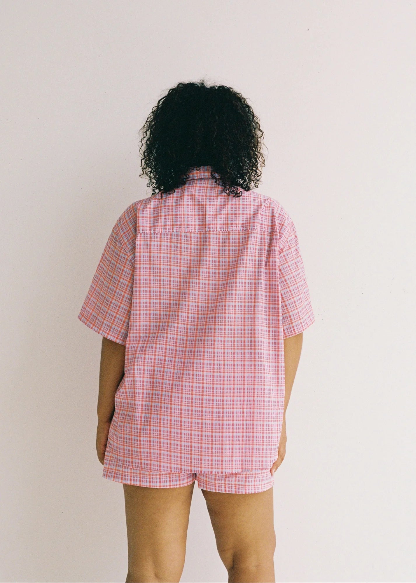 A person with curly hair is seen from behind, wearing an oversized fit of the JUNI Watson Shirt in a pink and blue checkered pattern. Made from 100% cotton, this short-sleeve shirt with matching shorts stands out against a plain white background.