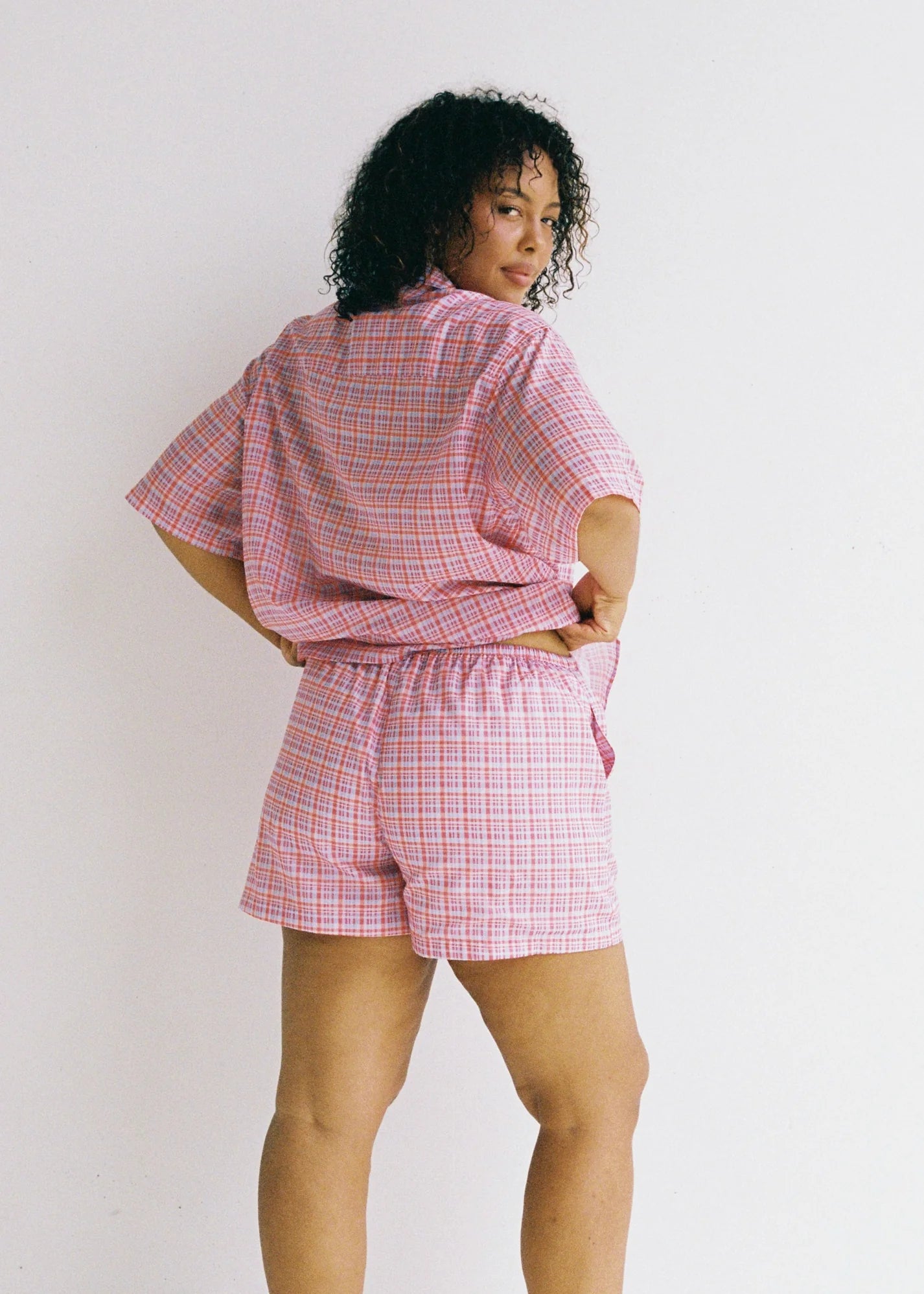 A person with curly hair looks over their shoulder, wearing JUNI's ethically made Dylan Short and matching pink and blue plaid shirt, both in a relaxed fit. The background is a plain white wall.