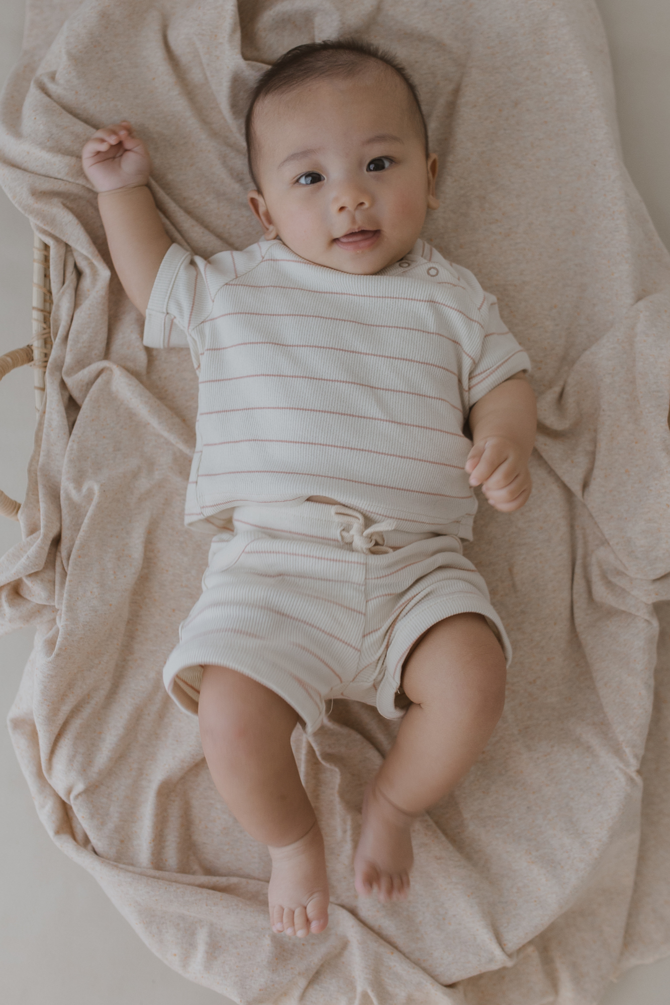 A baby dressed in the Boxy Tee Coco Stripe by SUSUKOSHI, made from organic cotton, rests on a soft beige blanket, gazing upwards with a gentle expression. One arm is slightly raised as the blanket is casually draped around them, creating a calm and cozy setting with the fabric's subtle relaxed fit.