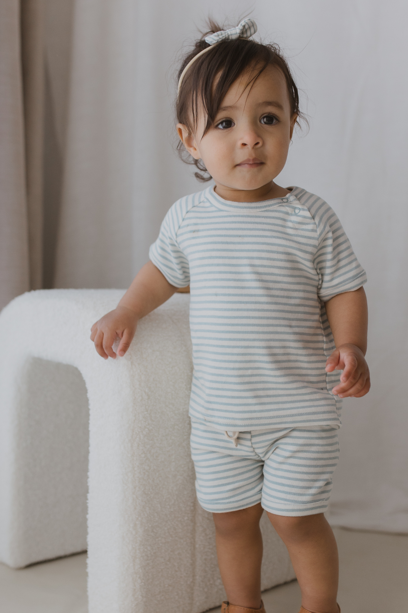 A toddler wearing the "Shorts Seaside" by SUSUKOSHI, paired with a striped outfit, stands beside a textured white chair. The child is accessorized with a headband featuring a bow and wears brown shoes, set against the backdrop of a soft, neutral curtain.