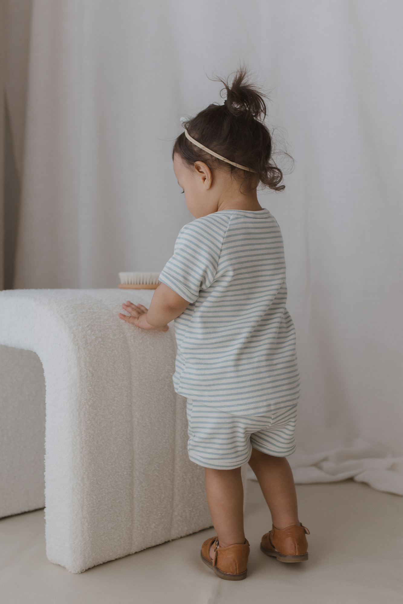 A toddler with dark hair in a small bun, wearing the "Shorts Seaside" from SUSUKOSHI and brown shoes, leans on a white textured piece of furniture. The toddler faces away, with a hairbrush visible on the furniture. The background has a neutral tone.