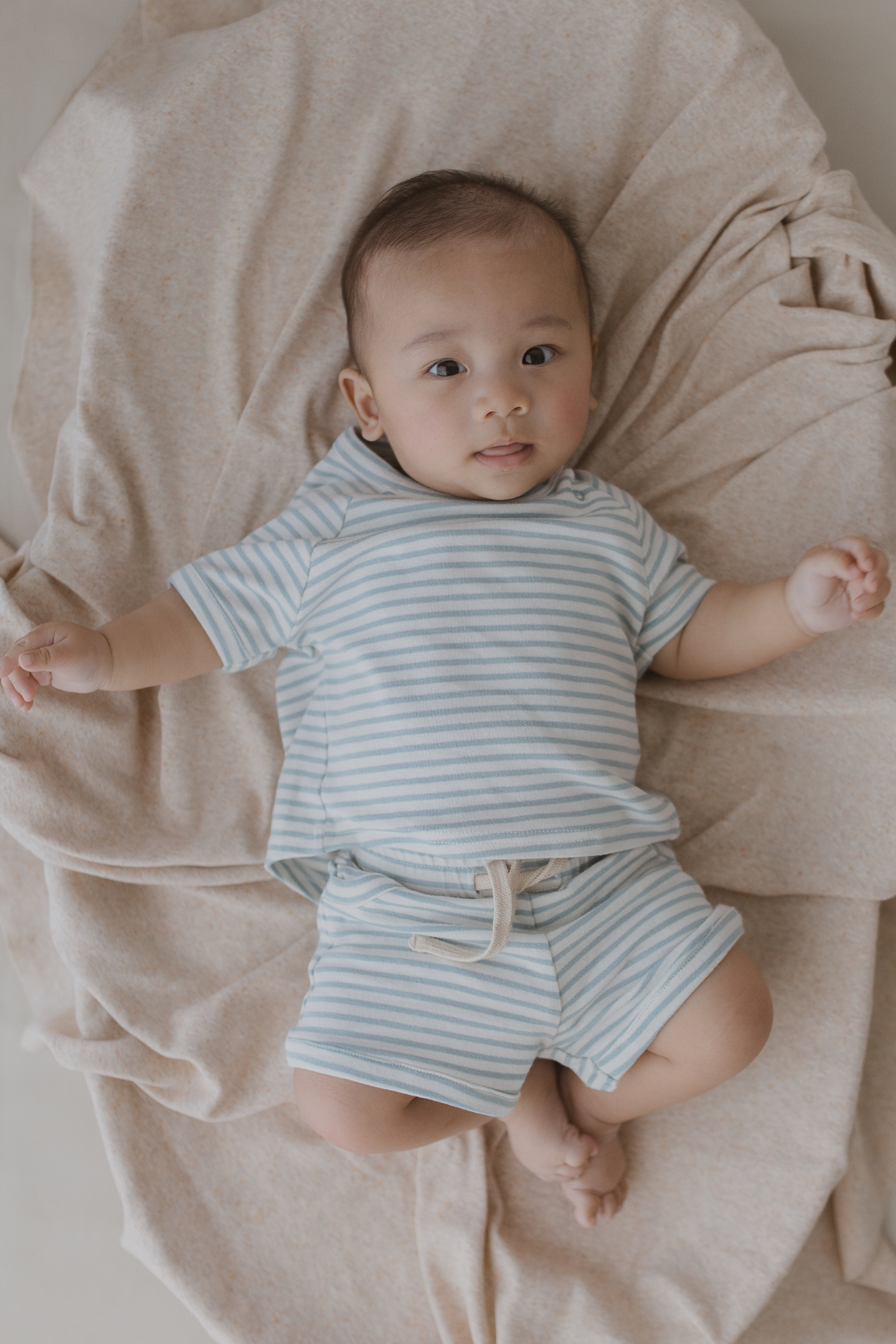 A baby with light brown hair is lying on a beige blanket, dressed in a light blue and white striped outfit, including the "Shorts Seaside" by SUSUKOSHI, featuring organic fabric and an adjustable drawstring. They gaze upwards with a relaxed expression, nestled among the soft folds of the fabric.