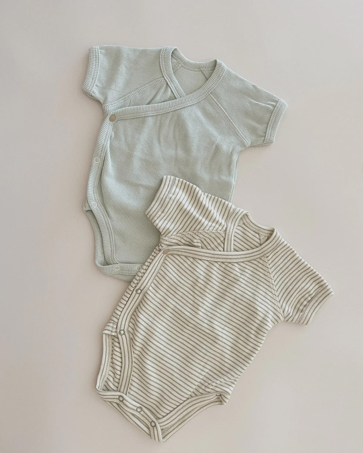 Two Kimono Bodysuits SS Sea Mist by SUSUKOSHI are laid flat on a creamy white surface. The top bodysuit, ideal for everyday wear, is solid light gray with a side snap closure, and the bottom one is white with thin gray stripes and a front snap closure. Both have short sleeves and are made from soft organic cotton elastane fabric.