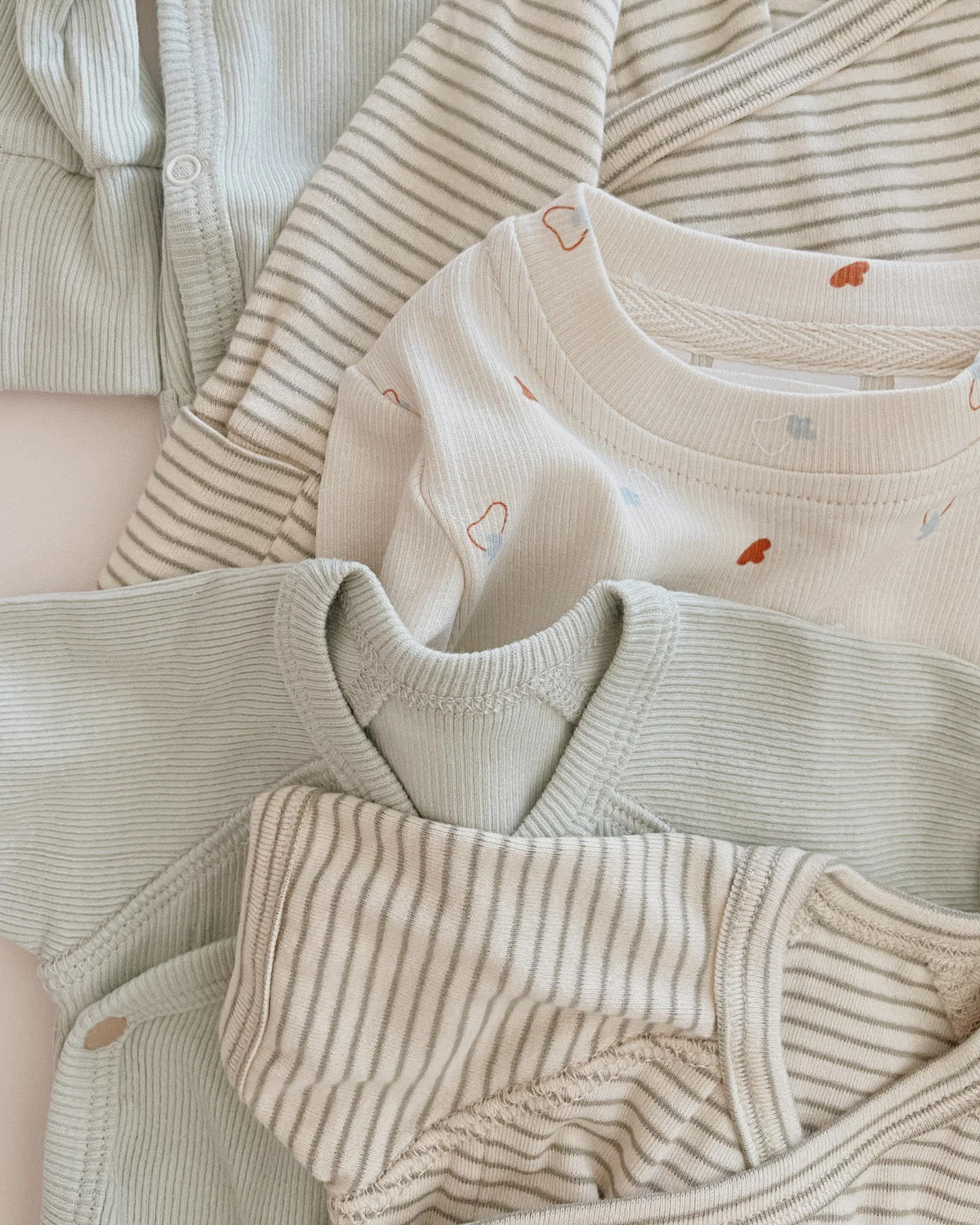 The Kimono Bodysuit SS Sea Mist by SUSUKOSHI is a collection of baby clothes in soft, neutral tones of white, beige, and light green. Featuring simple designs like stripes and small, discrete illustrations, these baby onesies are crafted from organic cotton elastane and neatly arranged in overlapping layers for display.