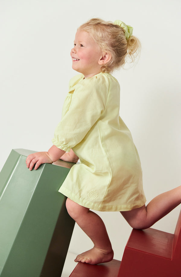 Blonde girl climbing on colourful stools wearing the Run to the Sun dress in a light fluro yellow colour