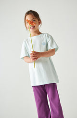 Girl in Rainbow Day tee and purple pants holding a flower in front of her face