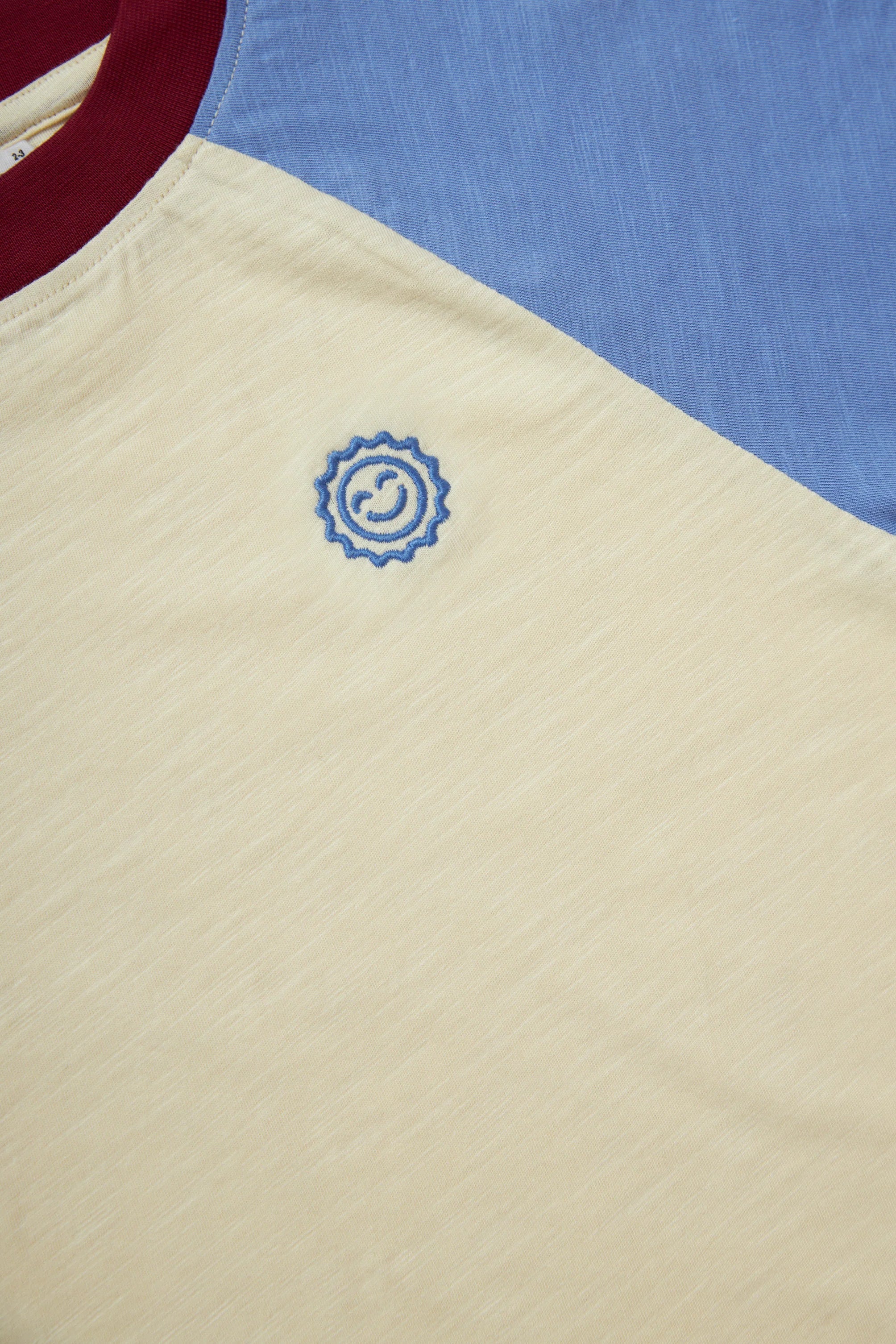 A close-up of KOKOMO's Weekend Raglan Long Sleeve Tee showcases its relaxed fit and color-block design. The T-shirt features a beige area with a small embroidered spiral logo, contrasted by a blue section. Crafted from organic cotton, the maroon neckline adds a splash of color to the minimalist design.