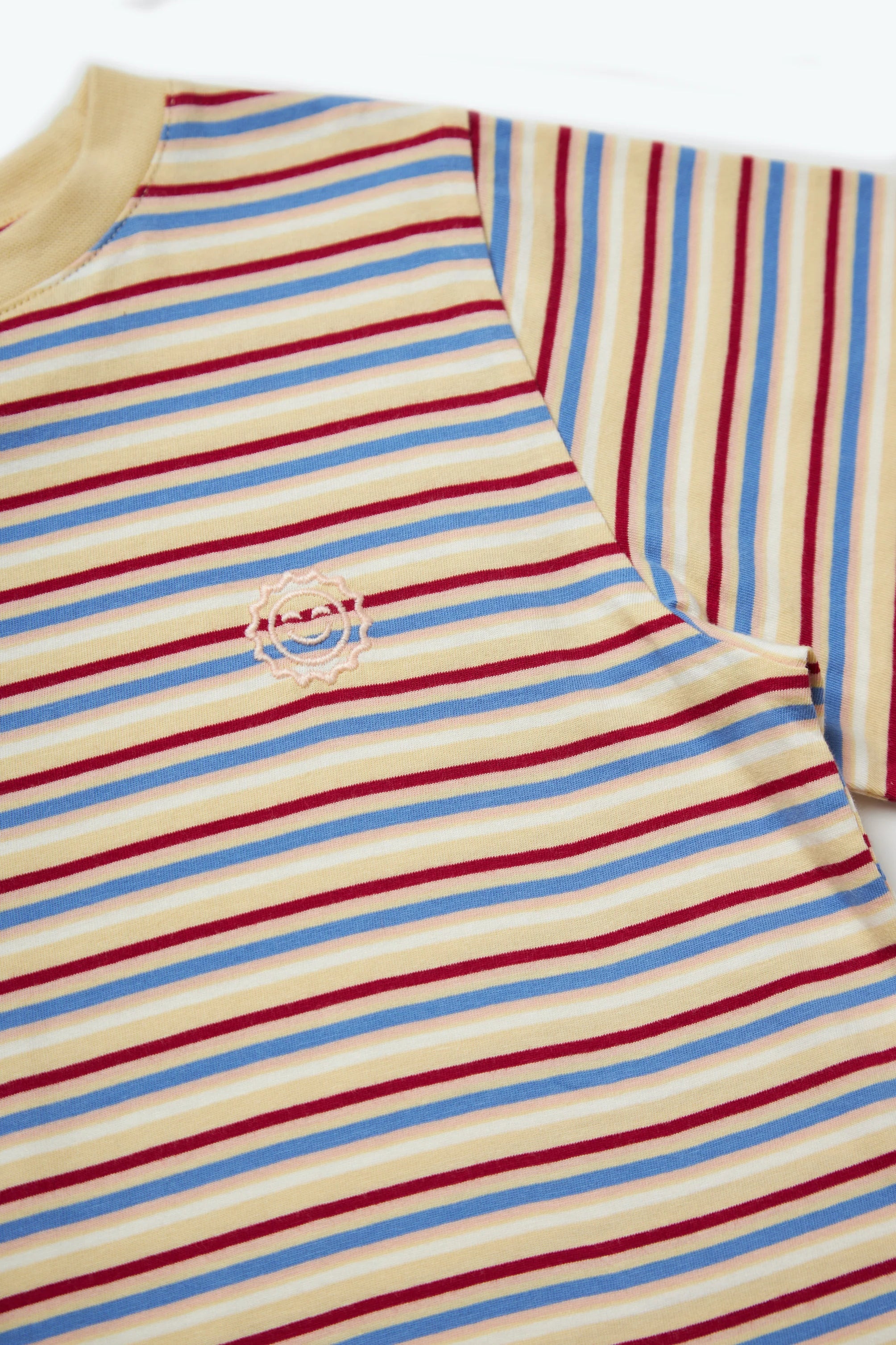 A close-up of the KOKOMO Stripe Sloppy Tee, featuring an oversized fit with horizontal red, blue, and beige stripes. This organic cotton shirt includes a circular embroidered logo near the chest area.