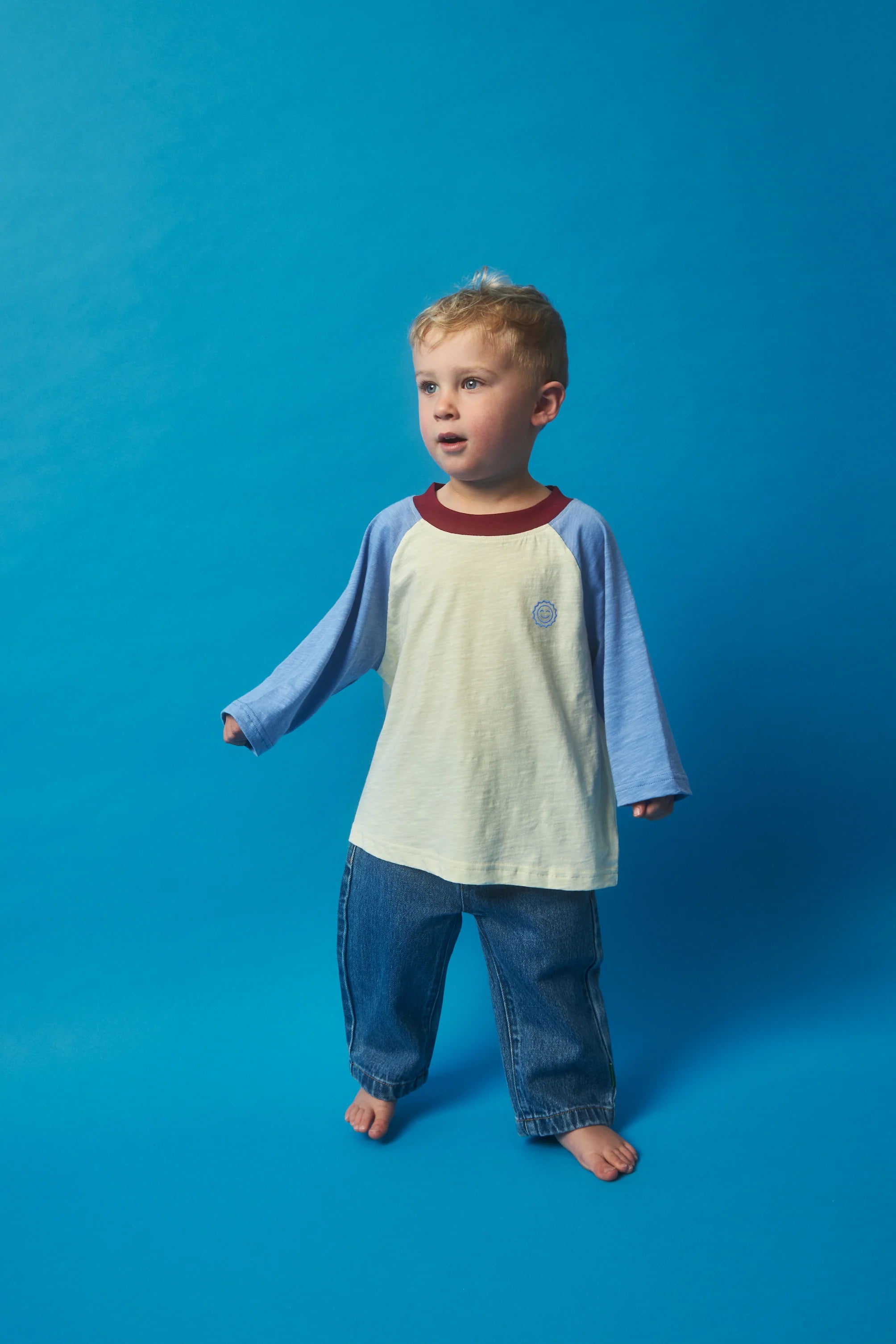 A young child stands barefoot against a blue background, wearing the KOKOMO Weekend Raglan Long Sleeve Tee, featuring blue raglan sleeves and a light-colored body, paired with jeans. The child's blond hair is slightly tousled.