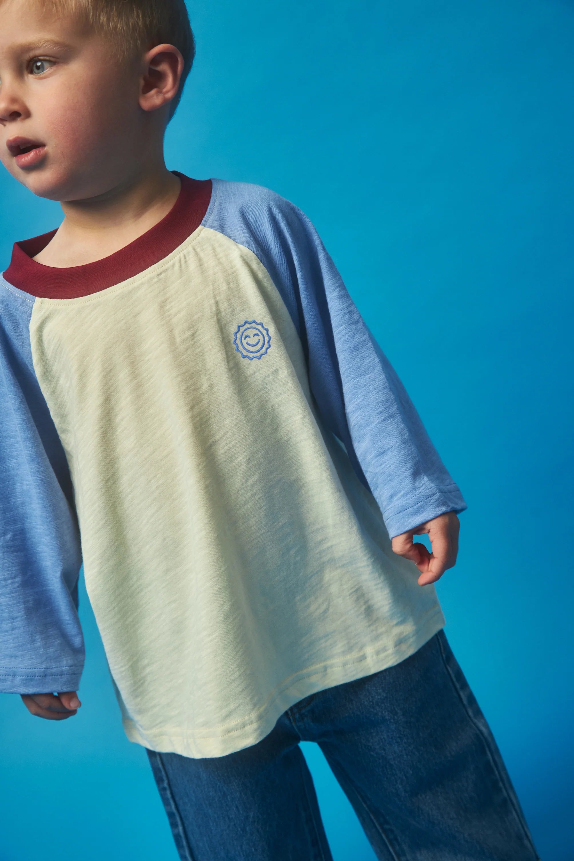 A young child is wearing the KOKOMO Weekend Raglan Long Sleeve Tee in light yellow and blue, crafted from organic cotton. The shirt showcases a small smiley face design, offering a relaxed fit with raglan sleeves for added comfort as the child stands against a blue background.