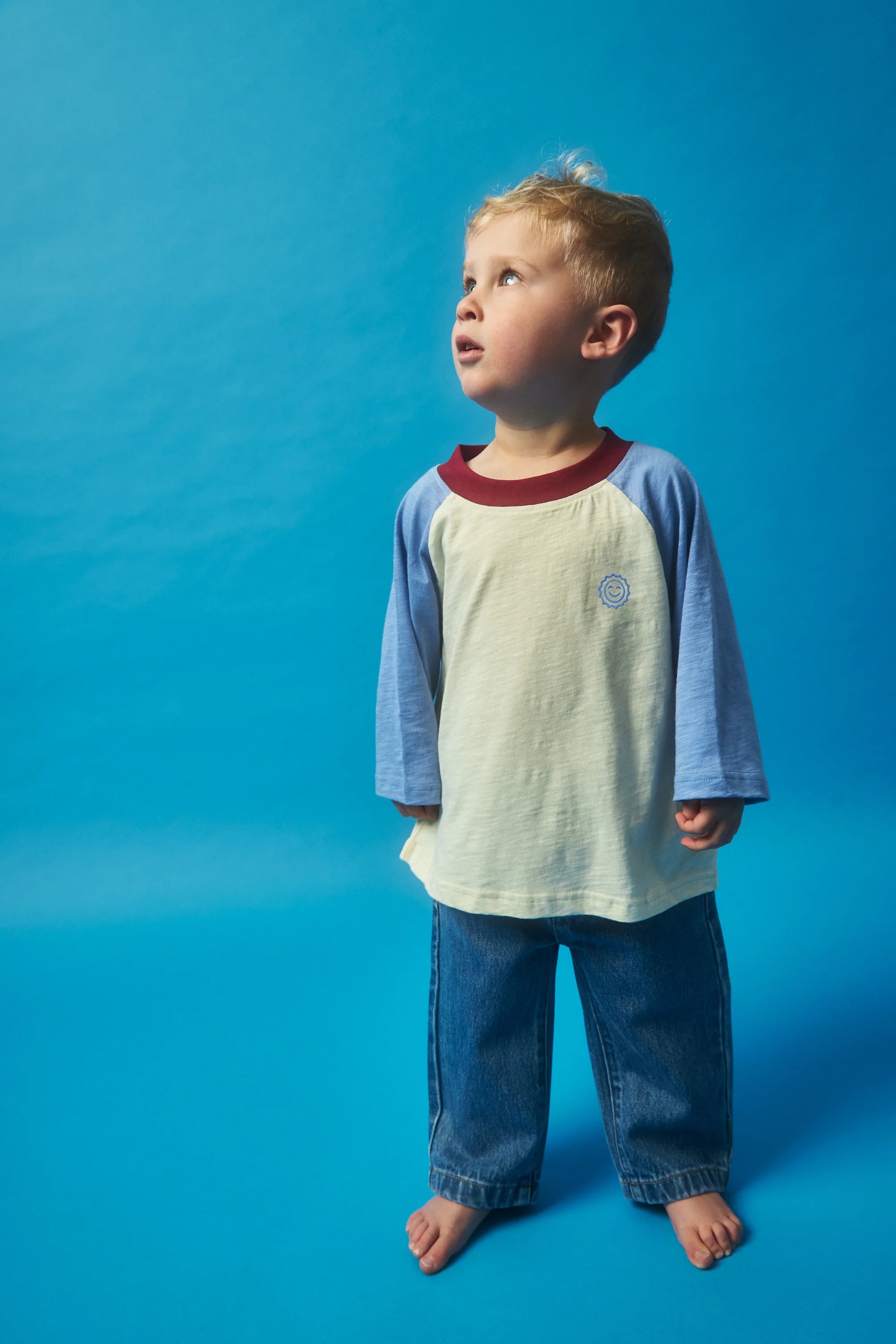 A young boy stands barefoot against a blue background, gazing upwards. He is dressed in KOKOMO's Weekend Raglan Long Sleeve Tee, featuring light blue raglan sleeves and a cream body made from organic cotton, complemented by relaxed fit blue jeans. His expression appears curious and thoughtful.