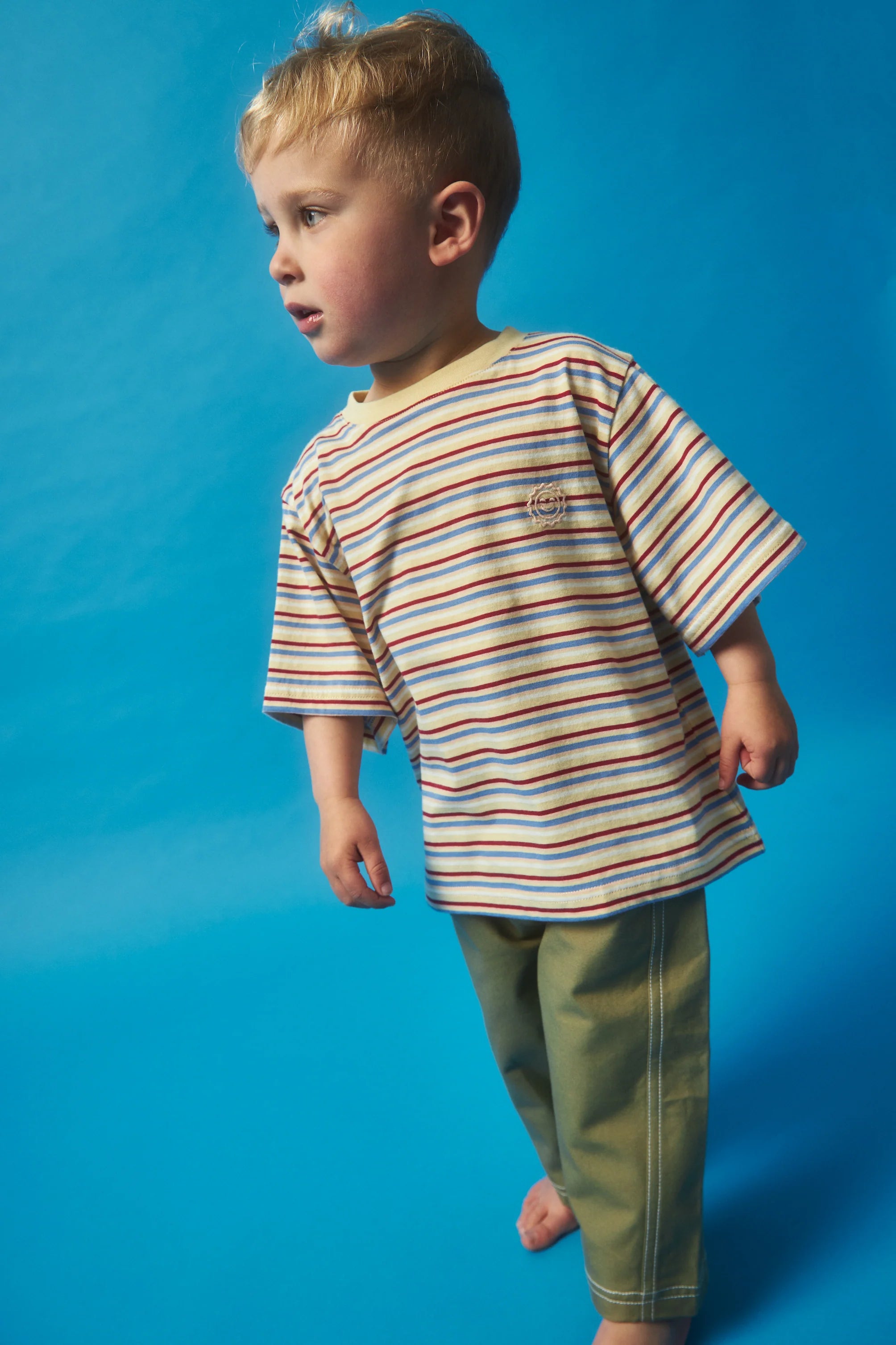 A young child wearing the Stripe Sloppy Tee by KOKOMO and green pants, both made from organic cotton for a soft touch, stands barefoot against a bright blue background, looking to the side. The child has short, light hair.