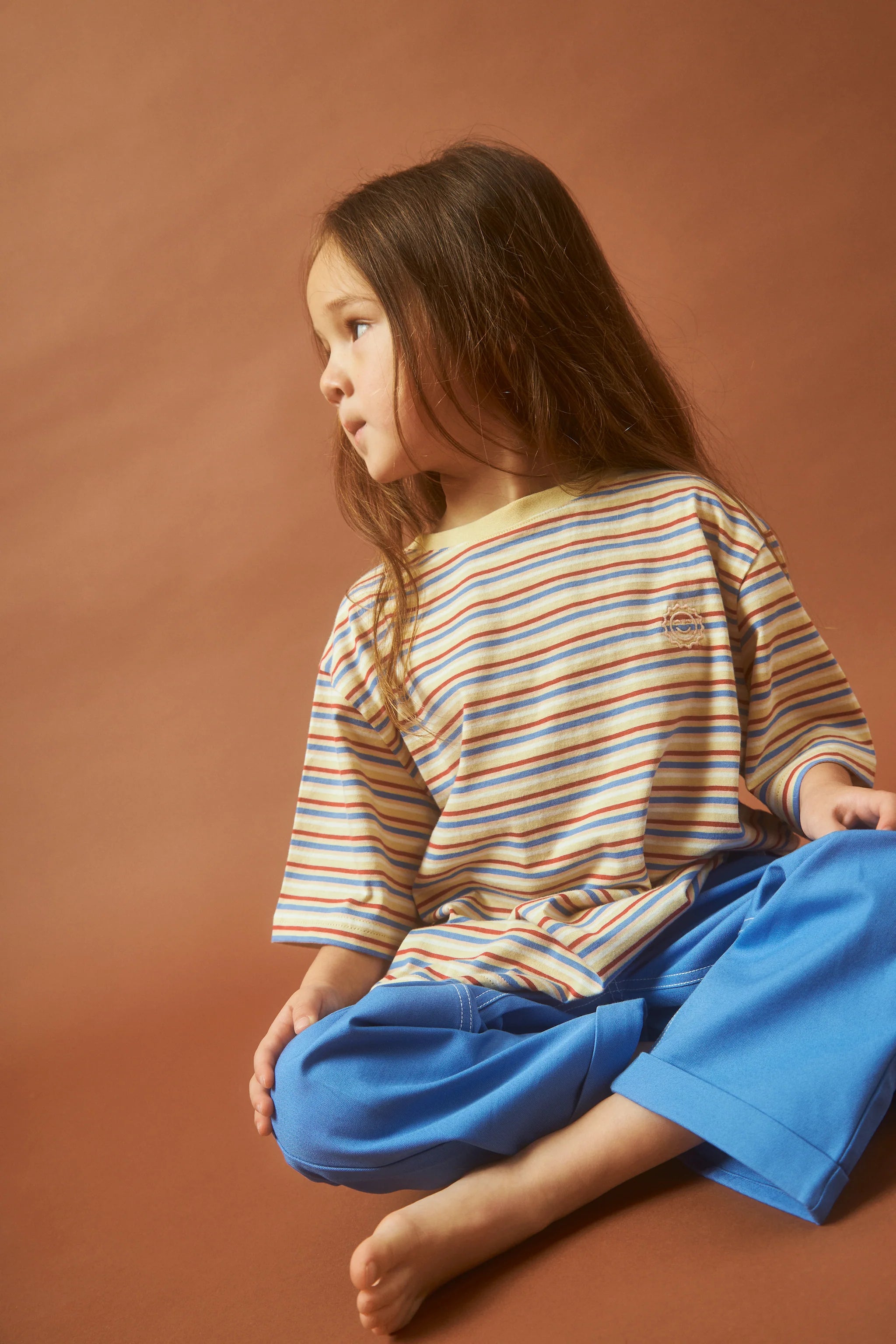 A young child with long hair is seated barefoot against a brown backdrop, wearing KOKOMO's Stripe Sloppy Tee made from organic cotton. The oversized shirt, featuring beige, blue, and orange stripes, pairs beautifully with their blue pants. With a thoughtful expression, they exude a peaceful aura that aligns with the Fairtrade Textile Standard.