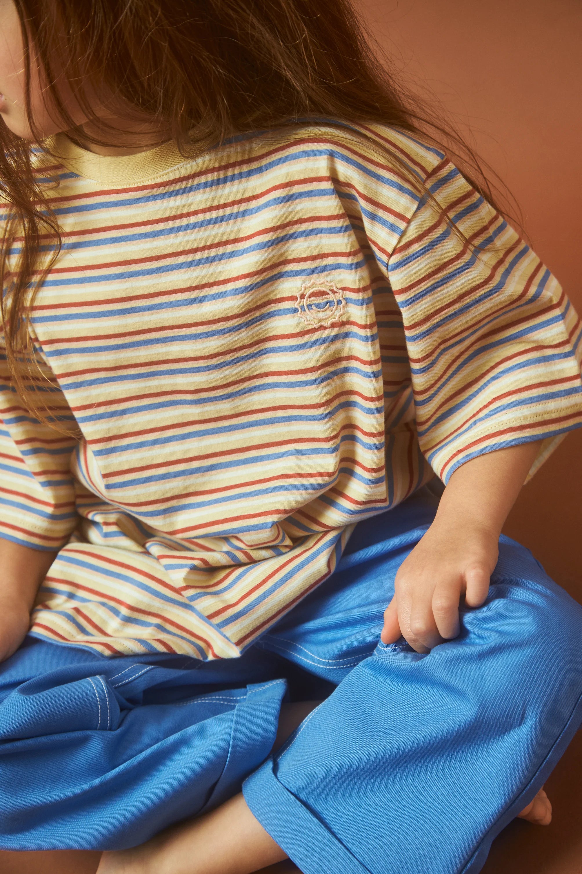 A child in an oversized Stripe Sloppy Tee by KOKOMO, featuring multicolored stripes and made from organic cotton, along with bright blue pants, sits cross-legged against a warm, earthy backdrop. The child's face remains out of frame in the image.