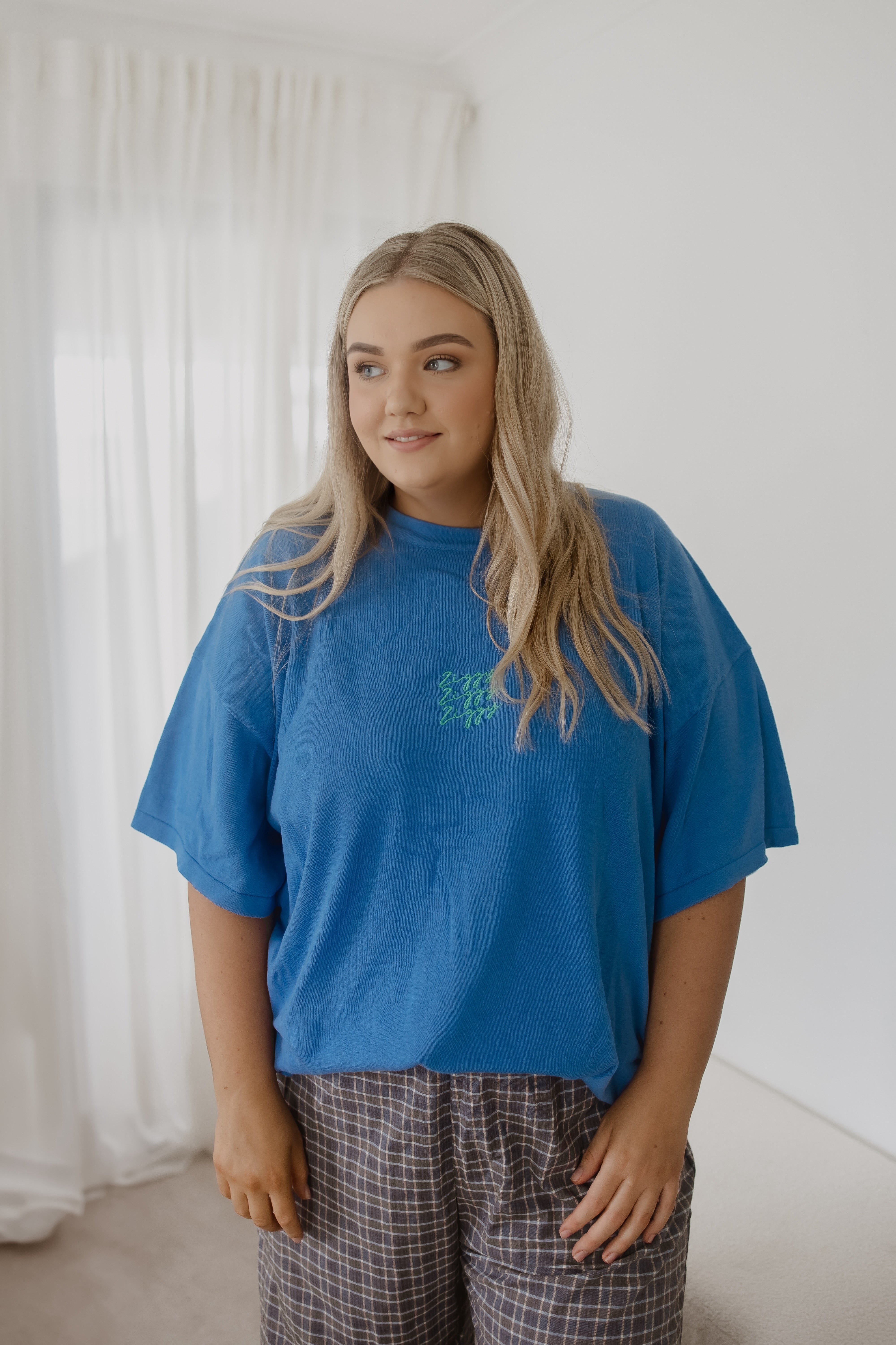 A young woman with long blonde hair stands indoors, wearing the Ziggy Lou - Women's Signature Tee Enzo by ZIGGY LOU and plaid pants. She smiles softly, looking to the side, with white curtains and light walls behind her.
