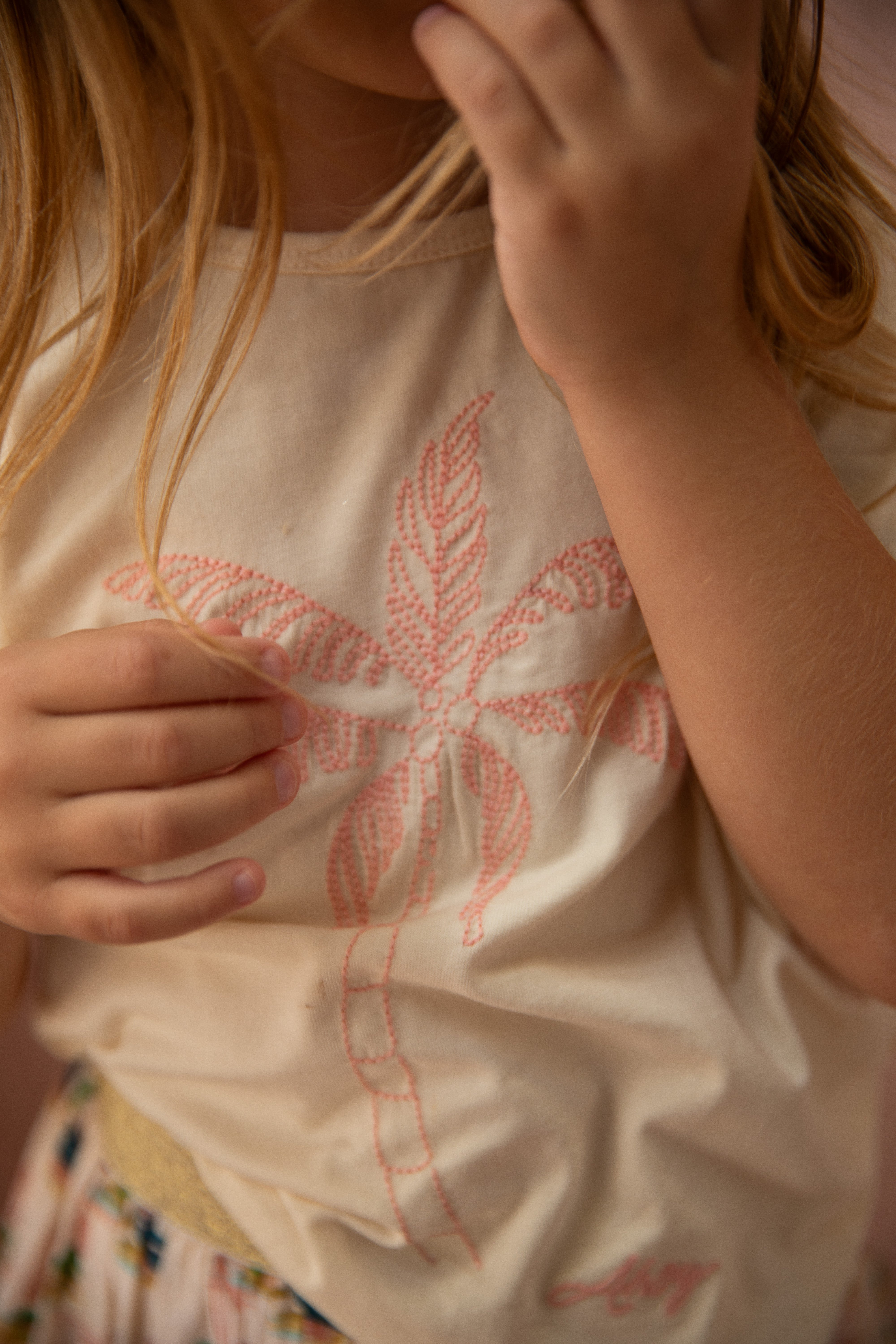 A child wearing the Key Largo Tee Vanilla by BELLA + LACE, featuring a beige cotton jersey material adorned with a pink palm tree design. The child's hand gently touches their shirt as their hair cascades over one shoulder. The softly blurred background encapsulates the essence of a sunny day ideal for enjoying ice cream.