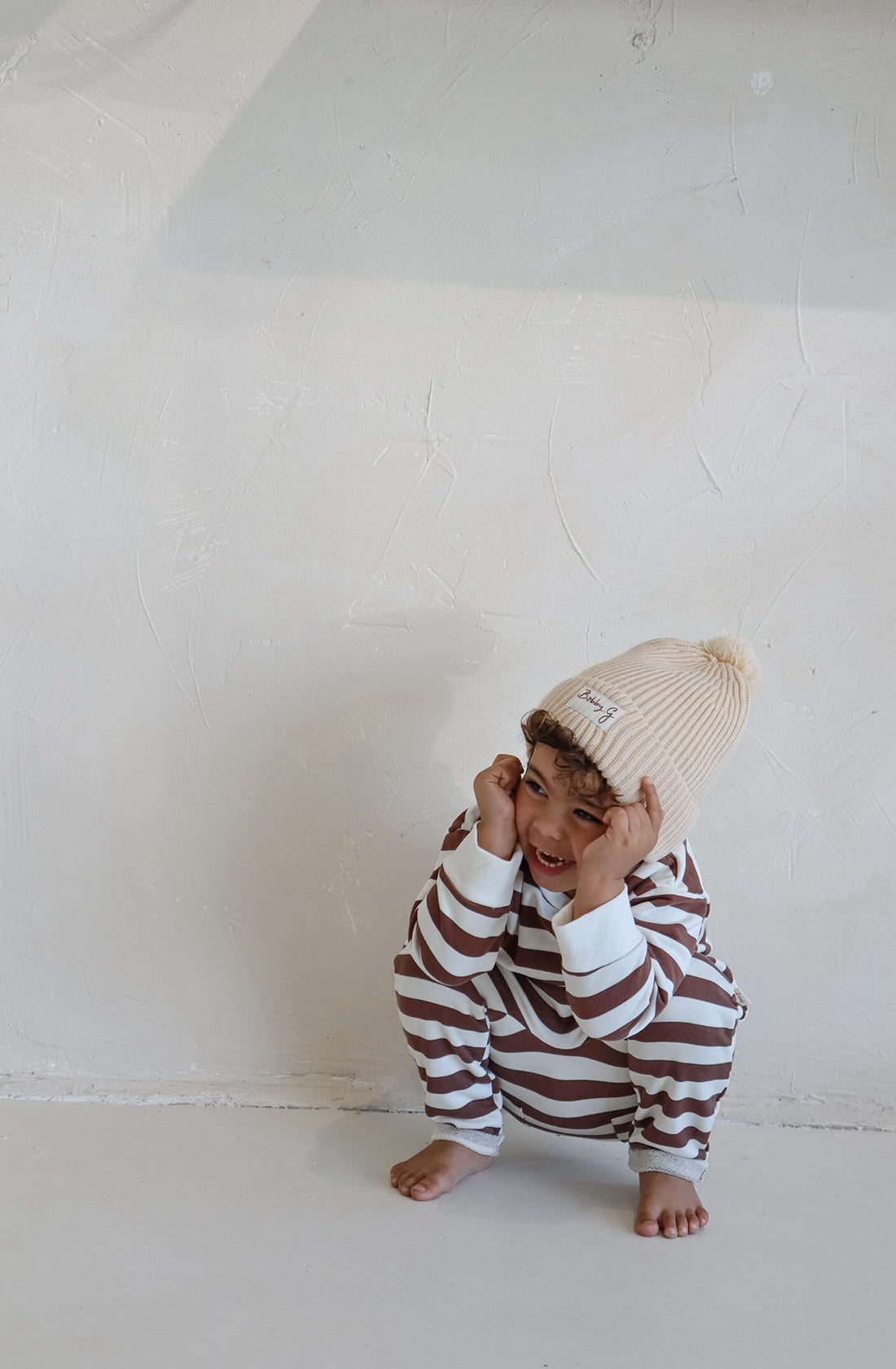 A young toddler wearing the sunny pant chocolate brown striped pants and matching pullover with the pom pom creme beanie.