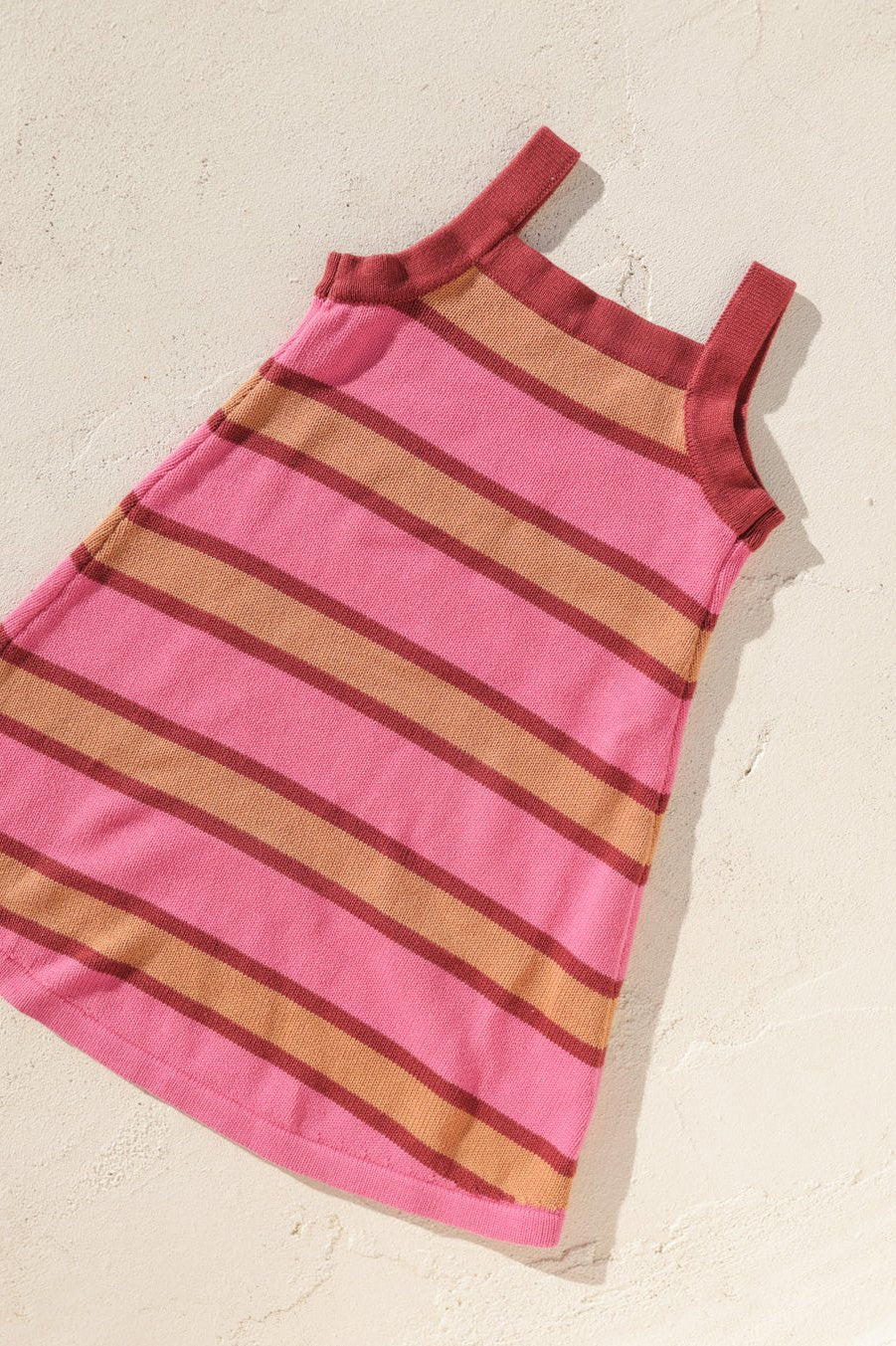 The Stripe Knit Dress by SMALL SWIM CLUB, featuring pink and brown horizontal stripes in a sleeveless A-line design, is displayed flat on a light-textured surface. Made from soft cotton, this dress offers everyday comfort with wide shoulder straps and a round neckline, ideal for effortless style.