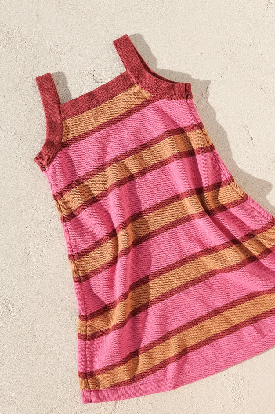 The Stripe Knit Dress by SMALL SWIM CLUB, featuring a pink and brown striped design, is laid flat on a light textured surface. This sleeveless cotton A-line dress displays horizontal stripes and wide straps, making it ideal for everyday wear.