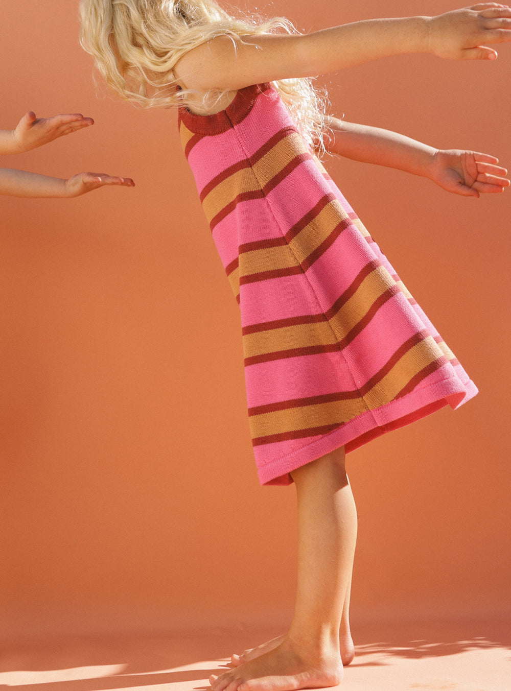 A child with long blonde hair, dressed in the Stripe Knit Dress from SMALL SWIM CLUB, reaches forward with outstretched arms against an orange background. Two hands approach the child from the side.