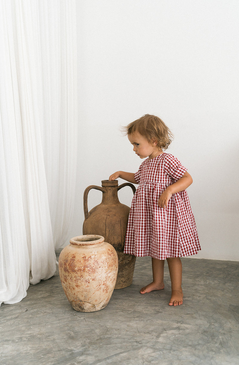 Toddler girl wearing red gingham dress standing next to two ceramic urns