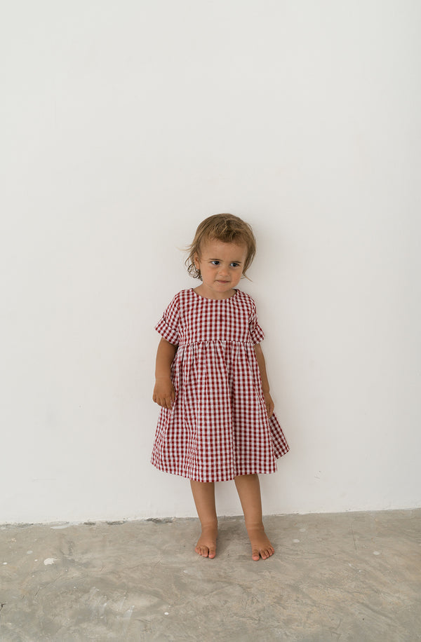Toddler girl wearing red gingham dress with bare feet standing against a white wall