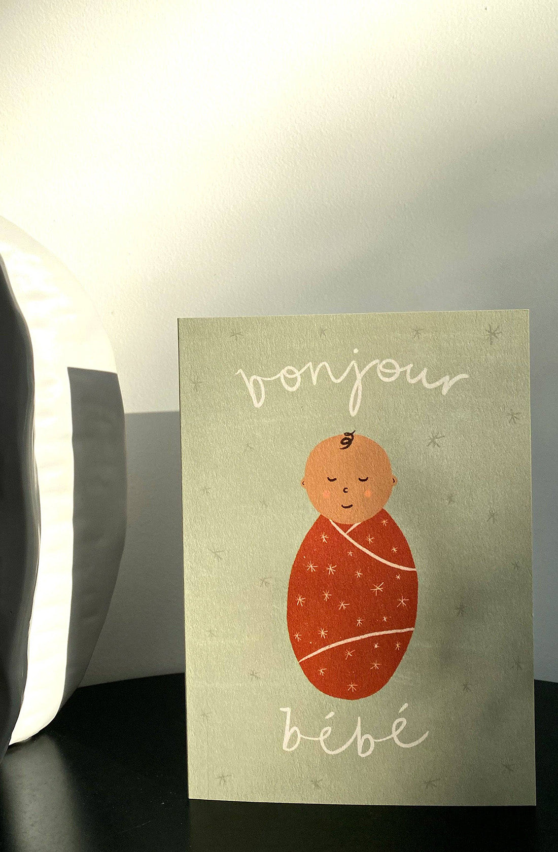 The Bonjour Bebe Card - New Baby by LAUREN SISSONS STUDIO features an illustration of a swaddled baby, complete with a small curl of hair, eyes closed, and a content expression. Set against a light green background, the card displays "bonjour bébé" in a playful font. This gender neutral greeting card is crafted from recycled paper and is designed to stand upright on any surface.