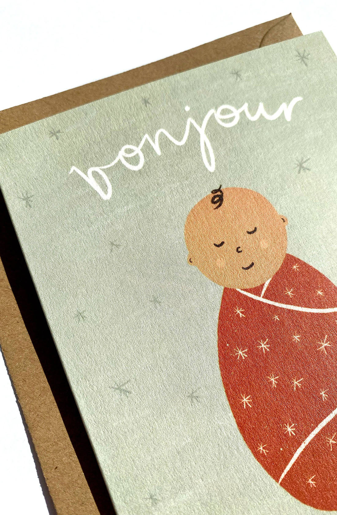 The Bonjour Bebe Card - New Baby by LAUREN SISSONS STUDIO features an illustrated baby swaddled in a red blanket with star patterns. The baby is set against a light green background, with the word "bonjour" beautifully written above in white cursive text. This gender-neutral greeting card is accompanied by a brown envelope made of recycled paper.