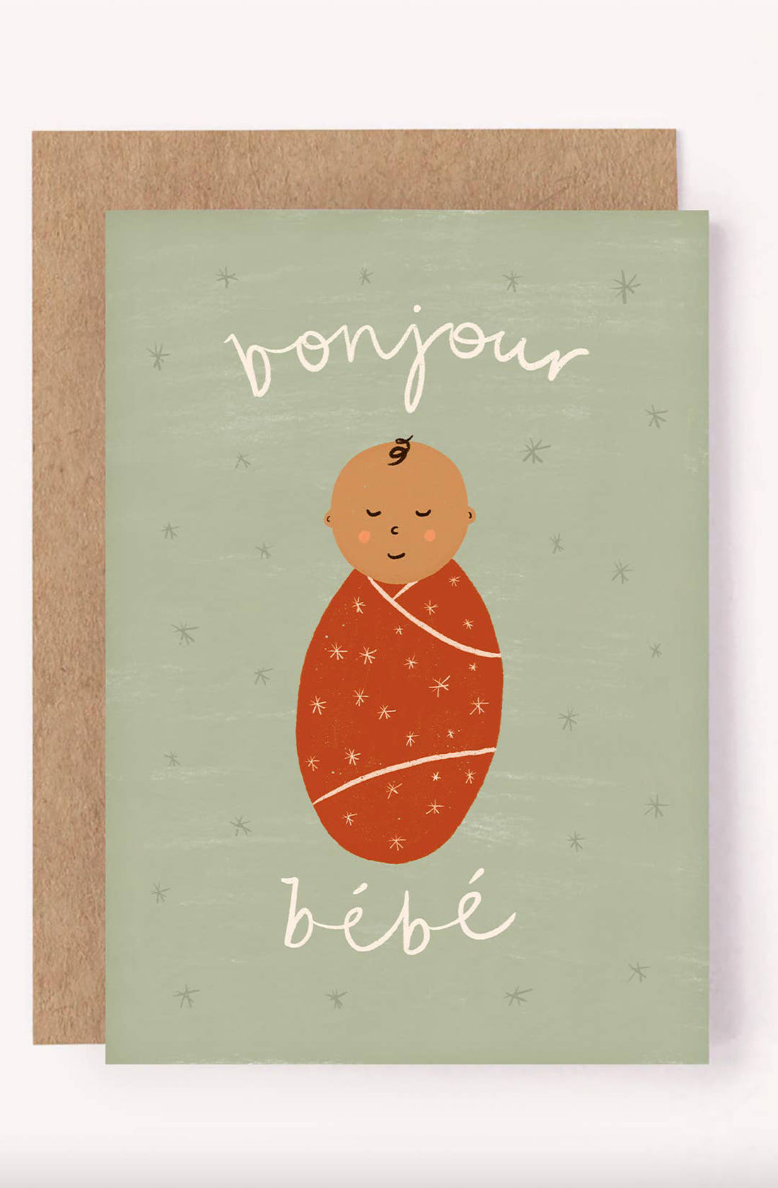 The Bonjour Bebe Card - New Baby by LAUREN SISSONS STUDIO is a gender-neutral greeting card with a light green background. It features an illustration of a swaddled baby in an orange blanket decorated with stars, with "Bonjour bébé" written above and below the baby in white cursive text. A brown envelope, crafted from recycled paper, peeks out from behind the card.