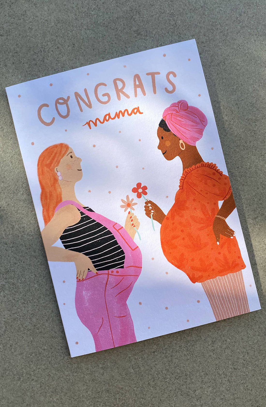 The Baby Shower Greeting Card "Congrats Mama" by LAUREN SISSONS STUDIO is perfect as a baby shower gift. It features an illustration of two pregnant women facing each other and smiling. One woman, with light skin and red hair, wears a striped top and pink overalls, while the other, with dark skin and a pink headwrap, is clad in a red dress. The text reads, "Congrats Mama.